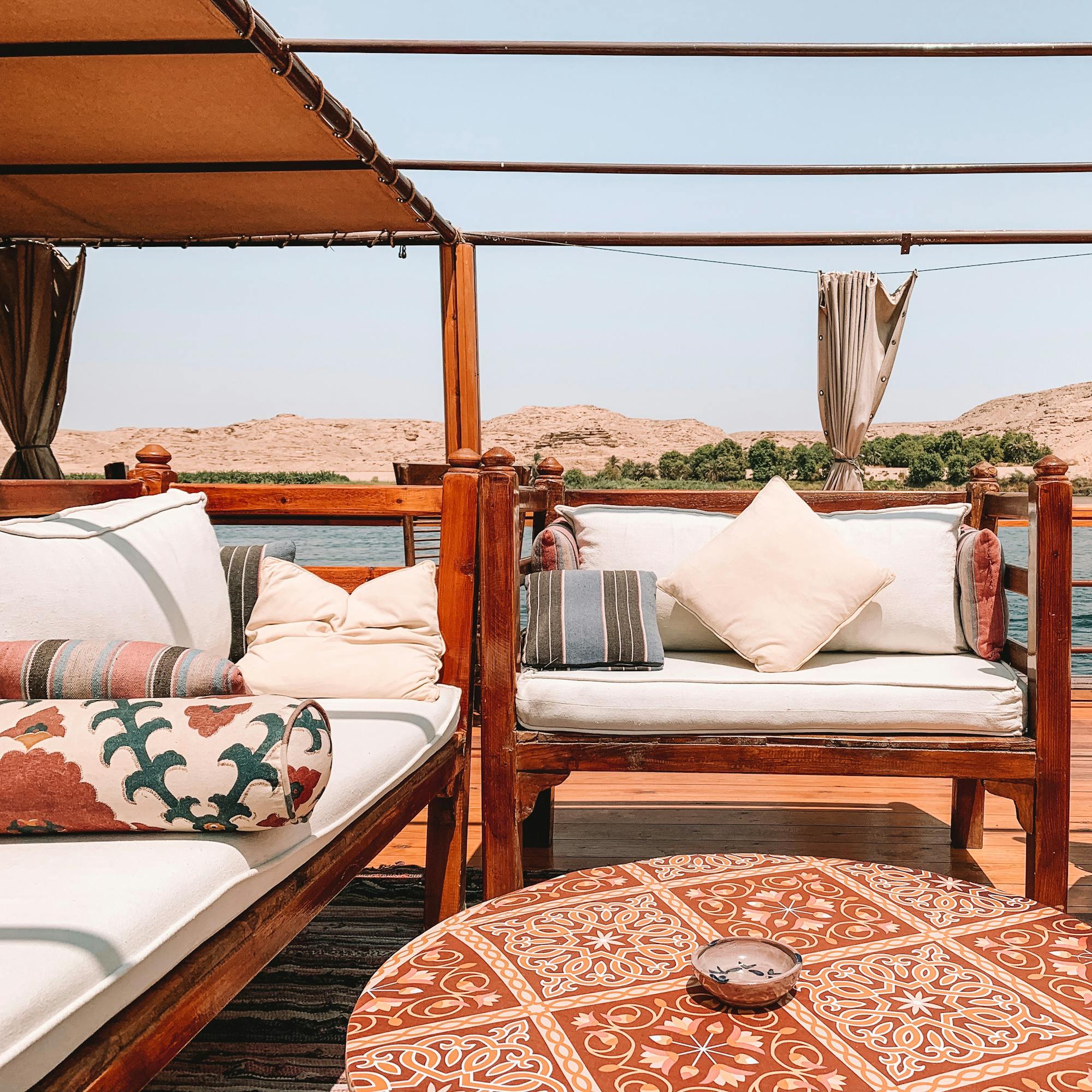 Daybed seating on a riverboat deck with cushions and canopy shade, looking out across the water and shoreline.