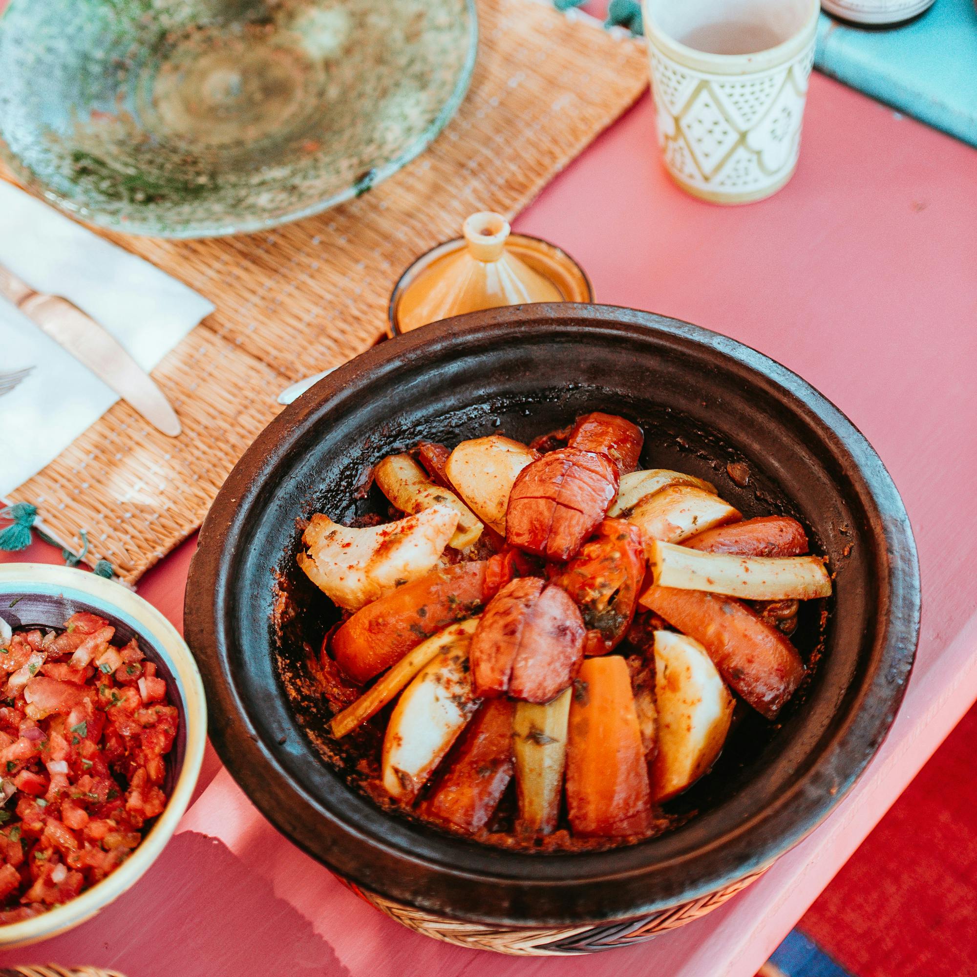 Colorful table spread with tagine, bread, and small dishes, arranged on patterned cloth with cups and bowls.