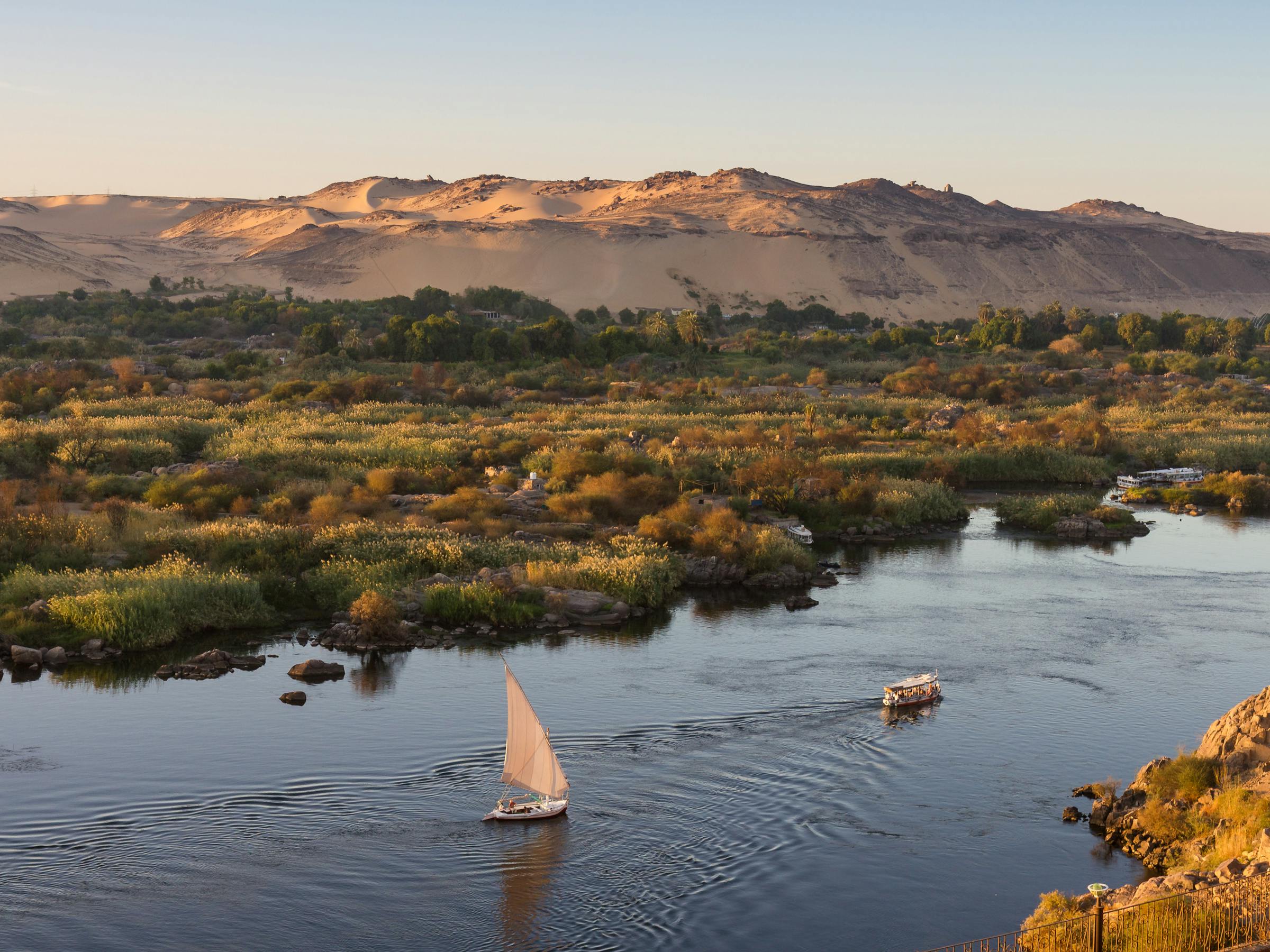 High view of a river with a small sailboat and green islands, as sunlight glints on the water at sunset below.