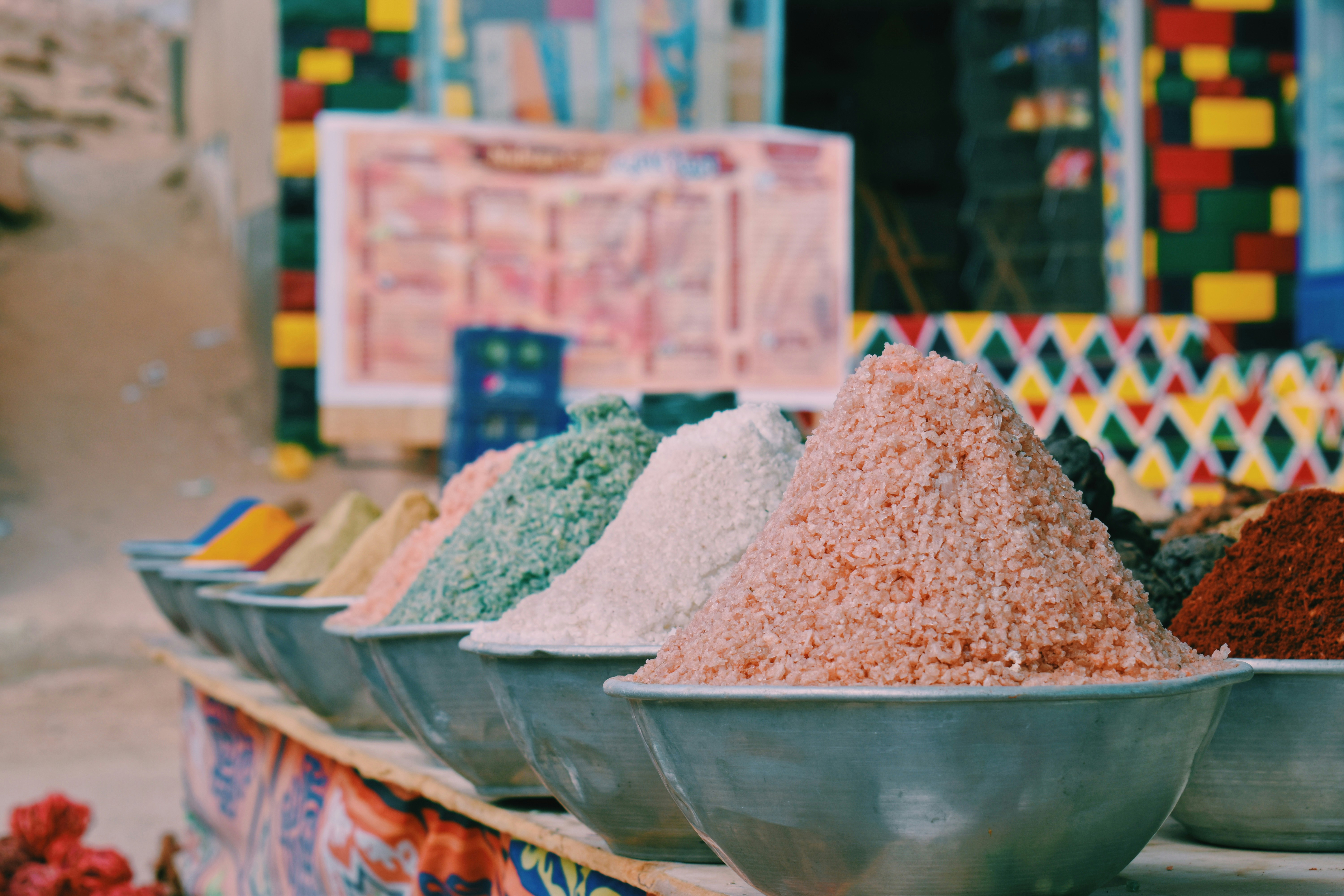 Large bowls of colorful spices fill a market stall, with patterned tiles and sacks arranged in the background.