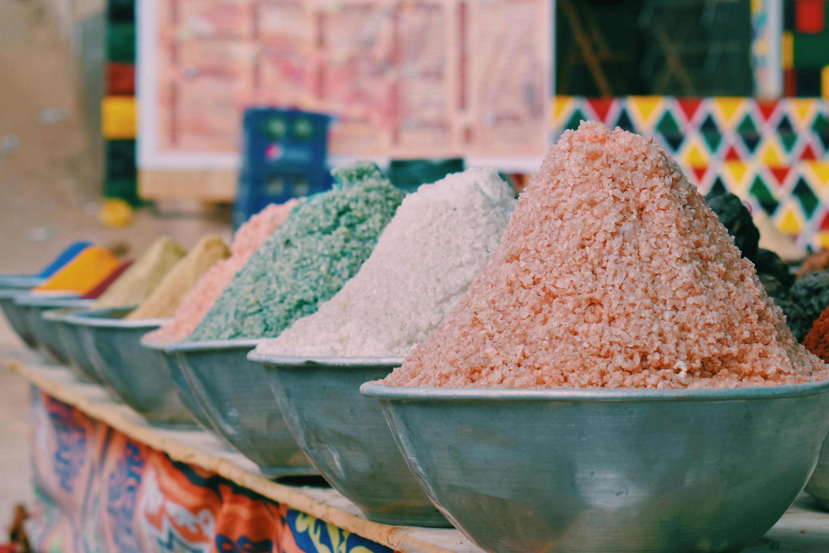 Large bowls of colorful spices fill a market stall, with patterned tiles and sacks arranged in the background.