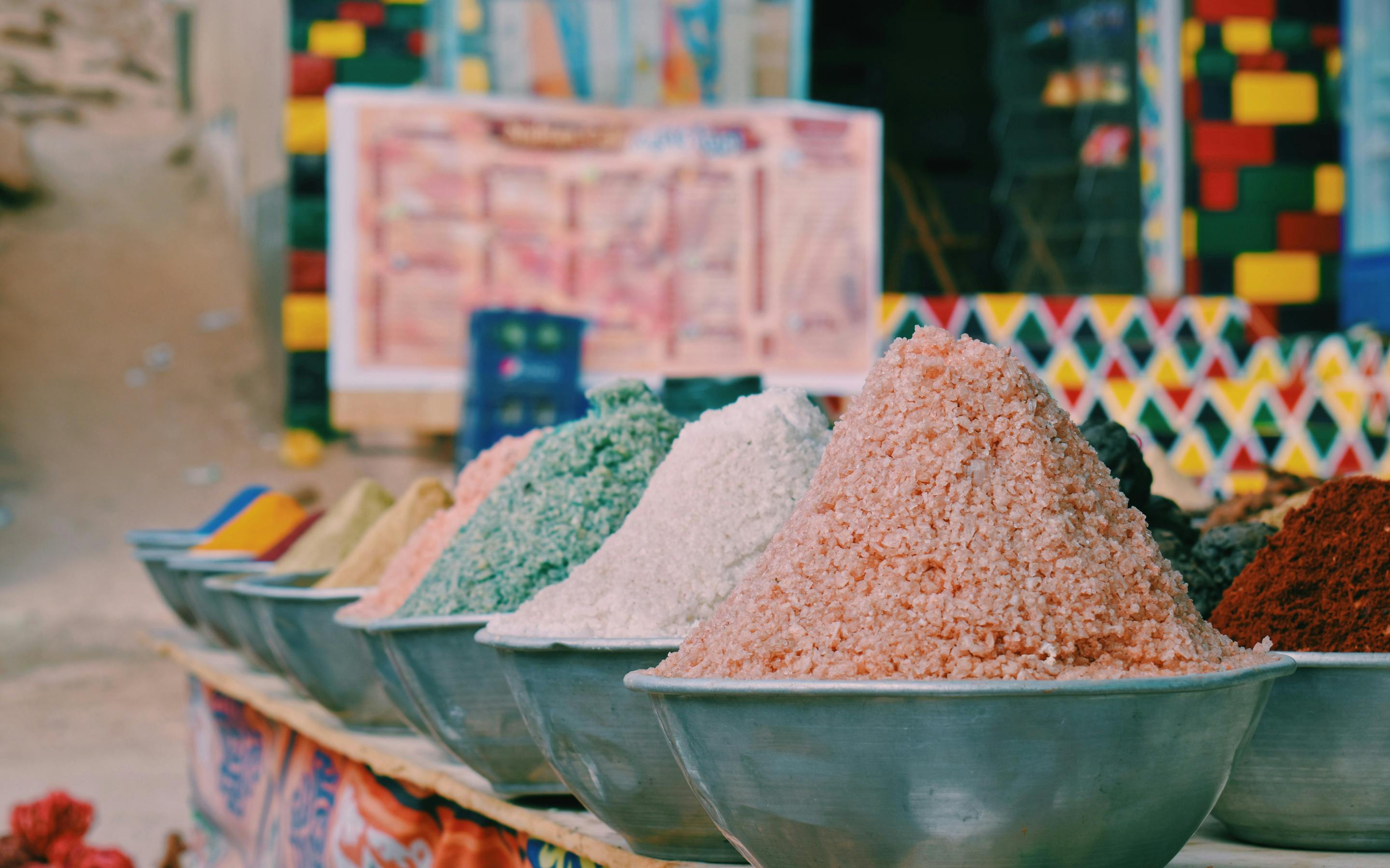 Large bowls of colorful spices fill a market stall, with patterned tiles and sacks arranged in the background.
