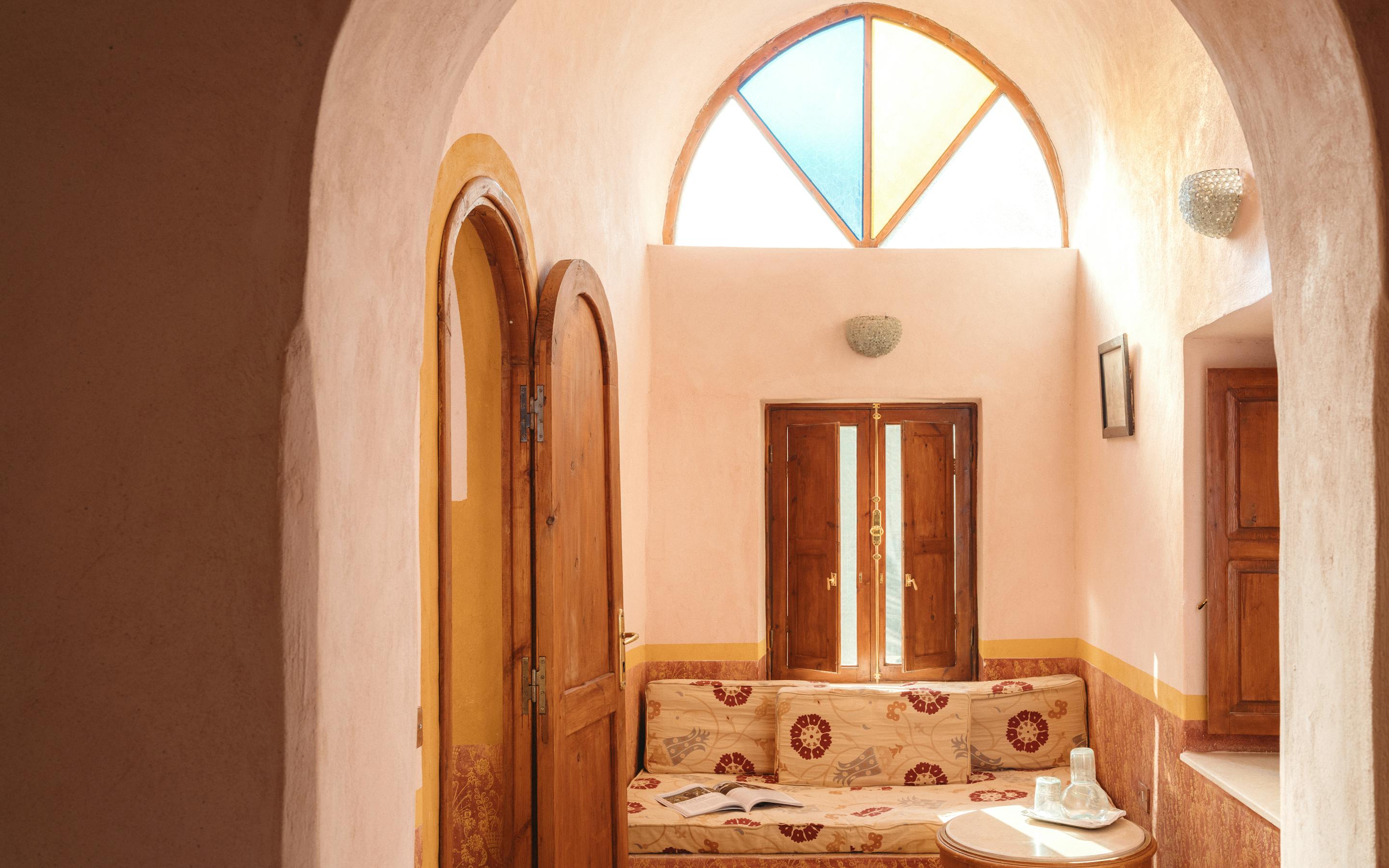 Sunlit arched doorway opens to a room with patterned tile floors and a single bed.