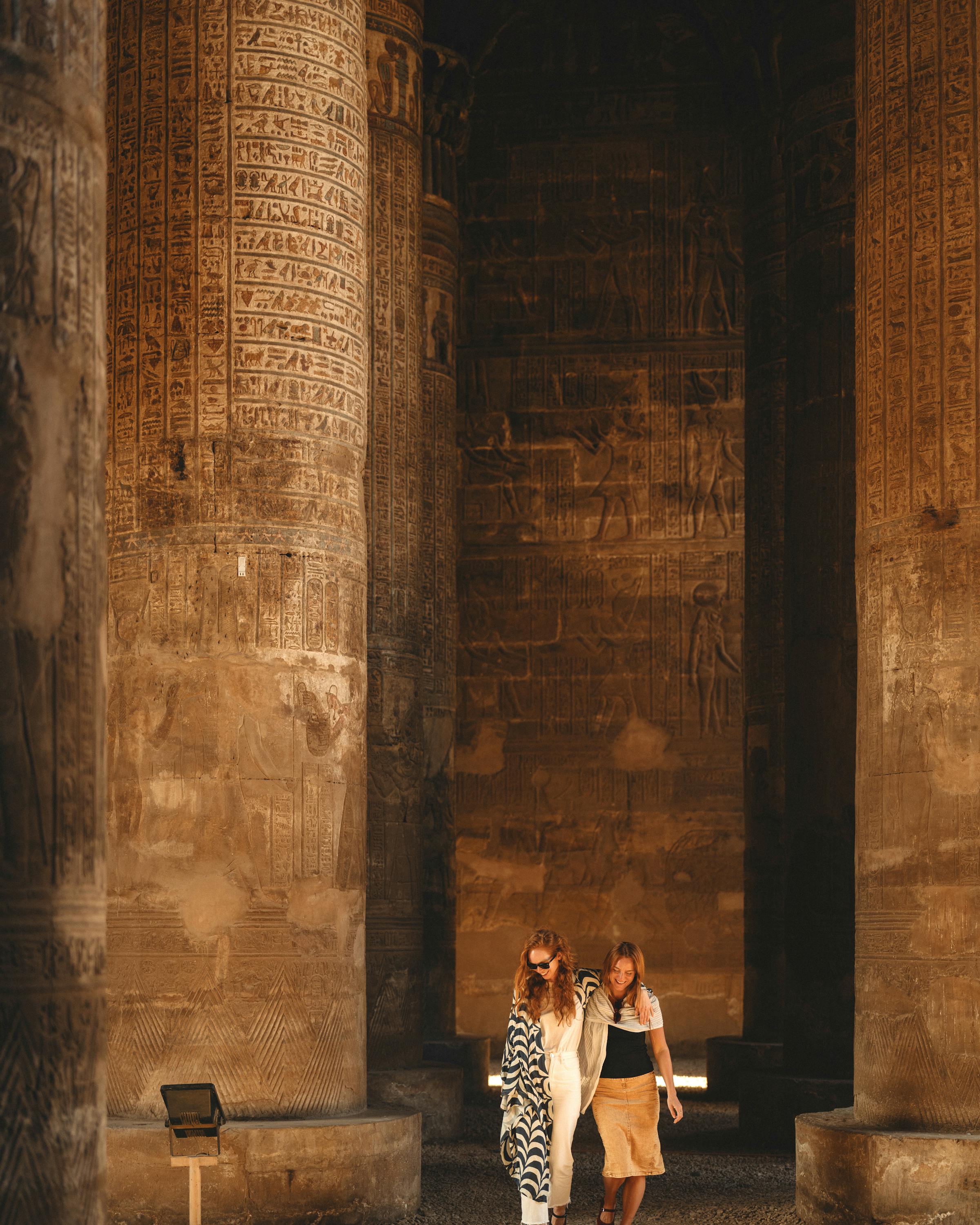 Two people walk between towering stone columns in a sunlit temple passage.
