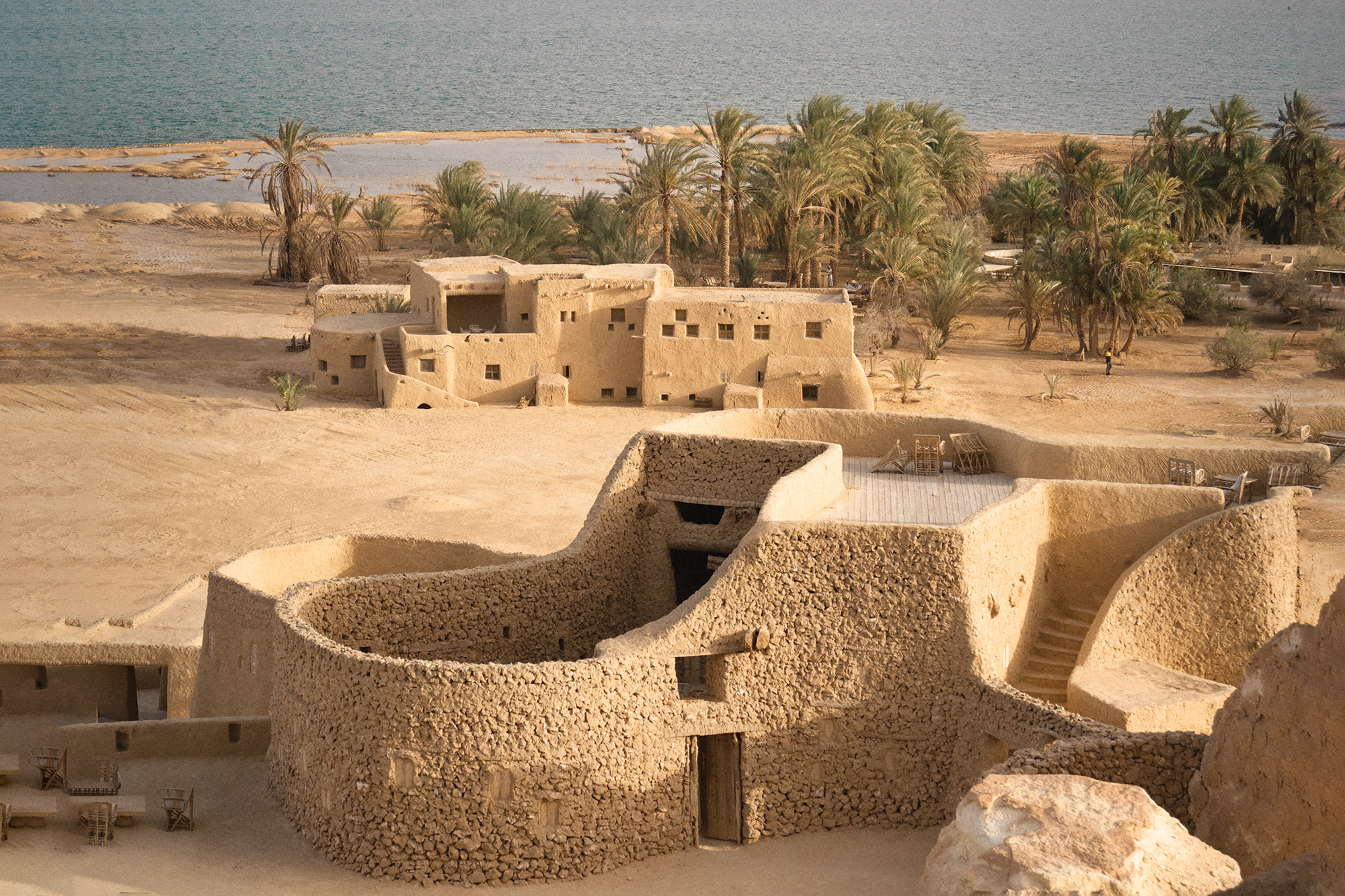 Sand-colored mudbrick buildings sit beside palm trees and water under a clear sky.