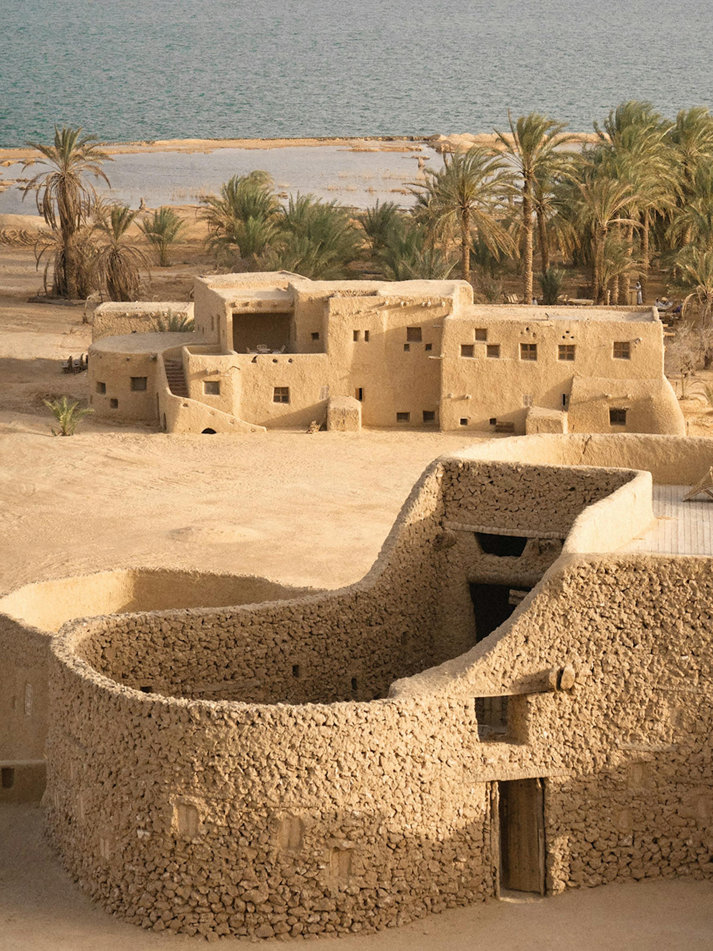 Sand-colored mudbrick buildings sit beside palm trees and water under a clear sky.