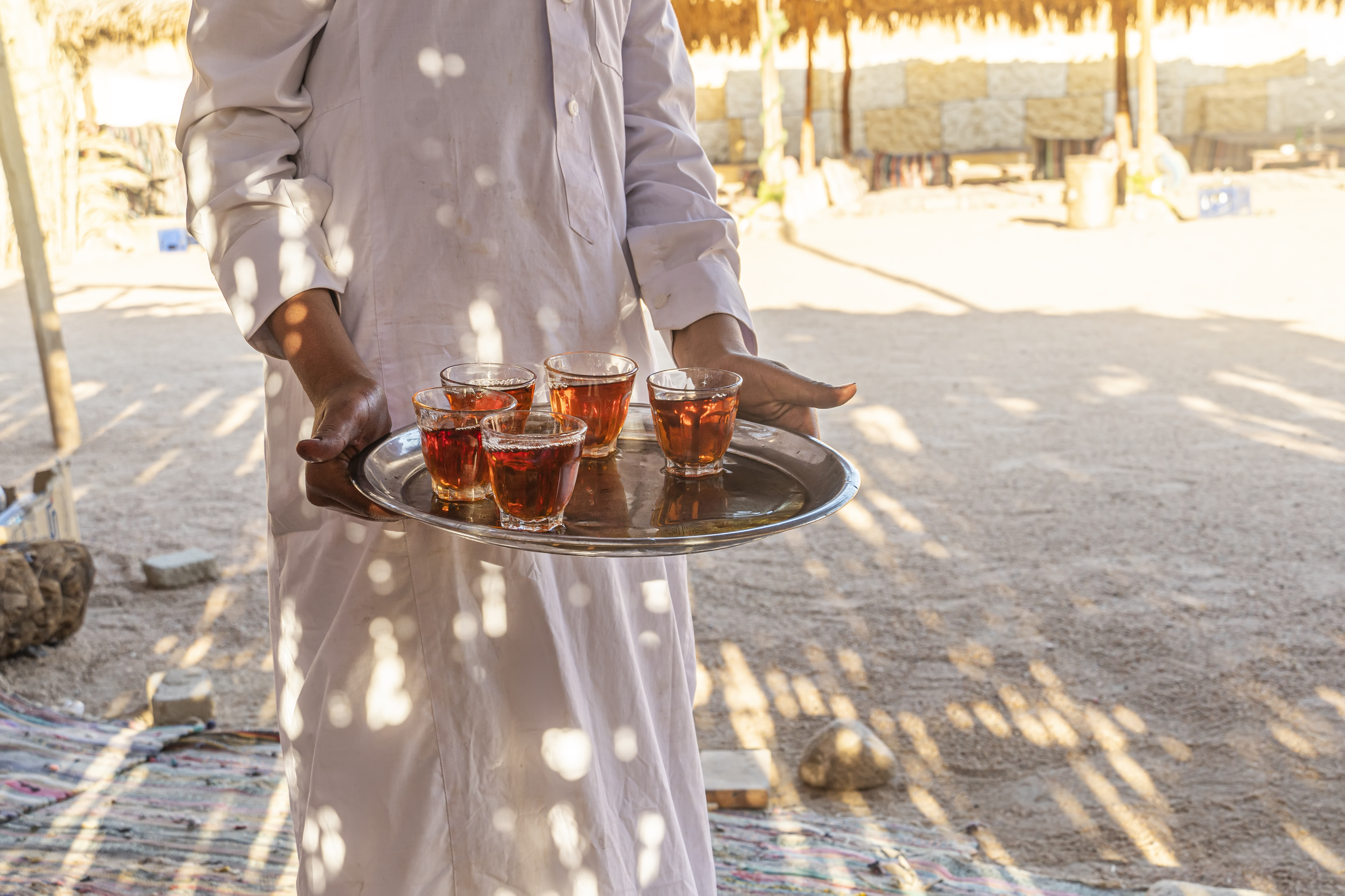A person carries a tray of small glasses on a sunlit deck with sand and dappled shadows.