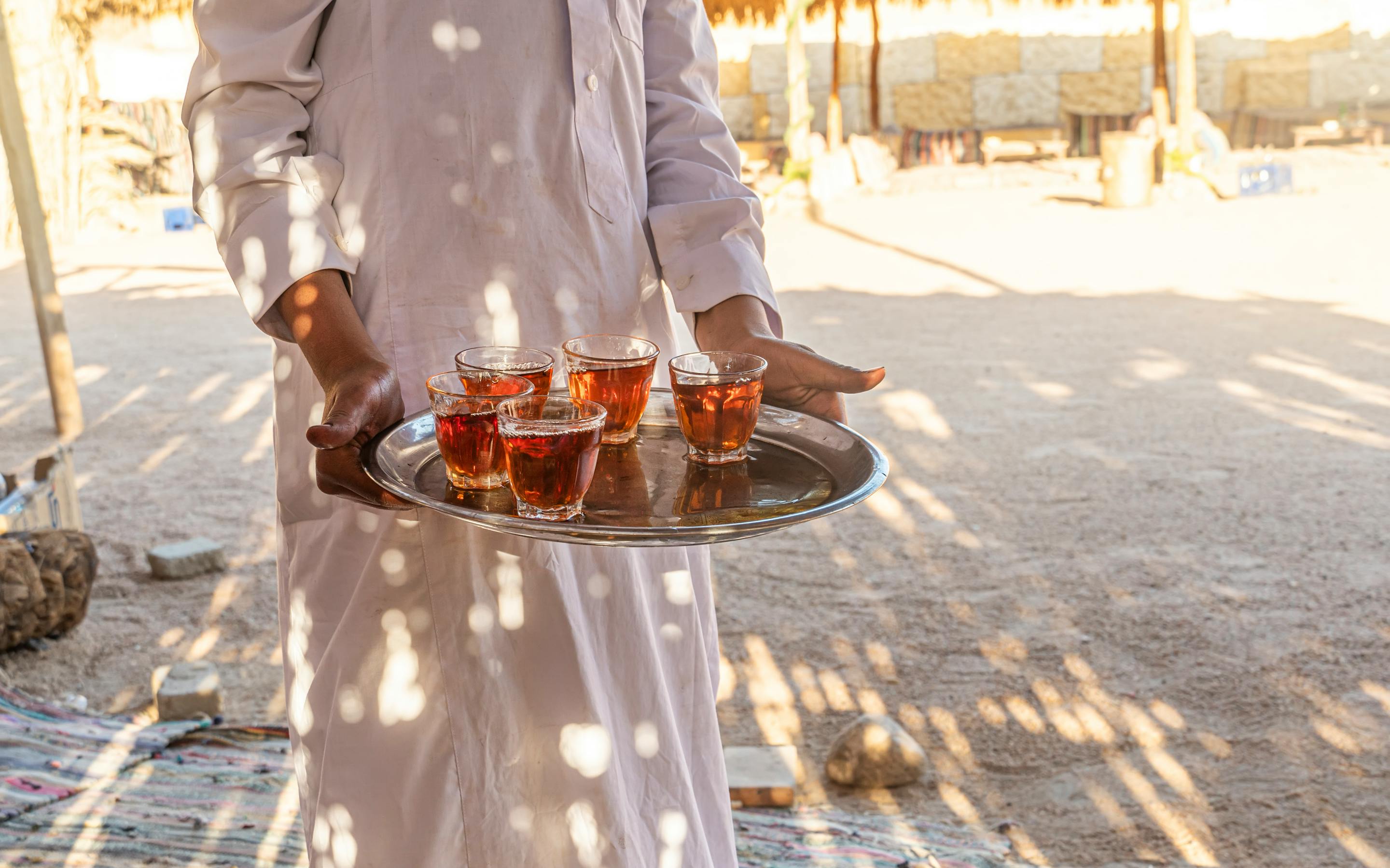 A person carries a tray of small glasses on a sunlit deck with sand and dappled shadows.