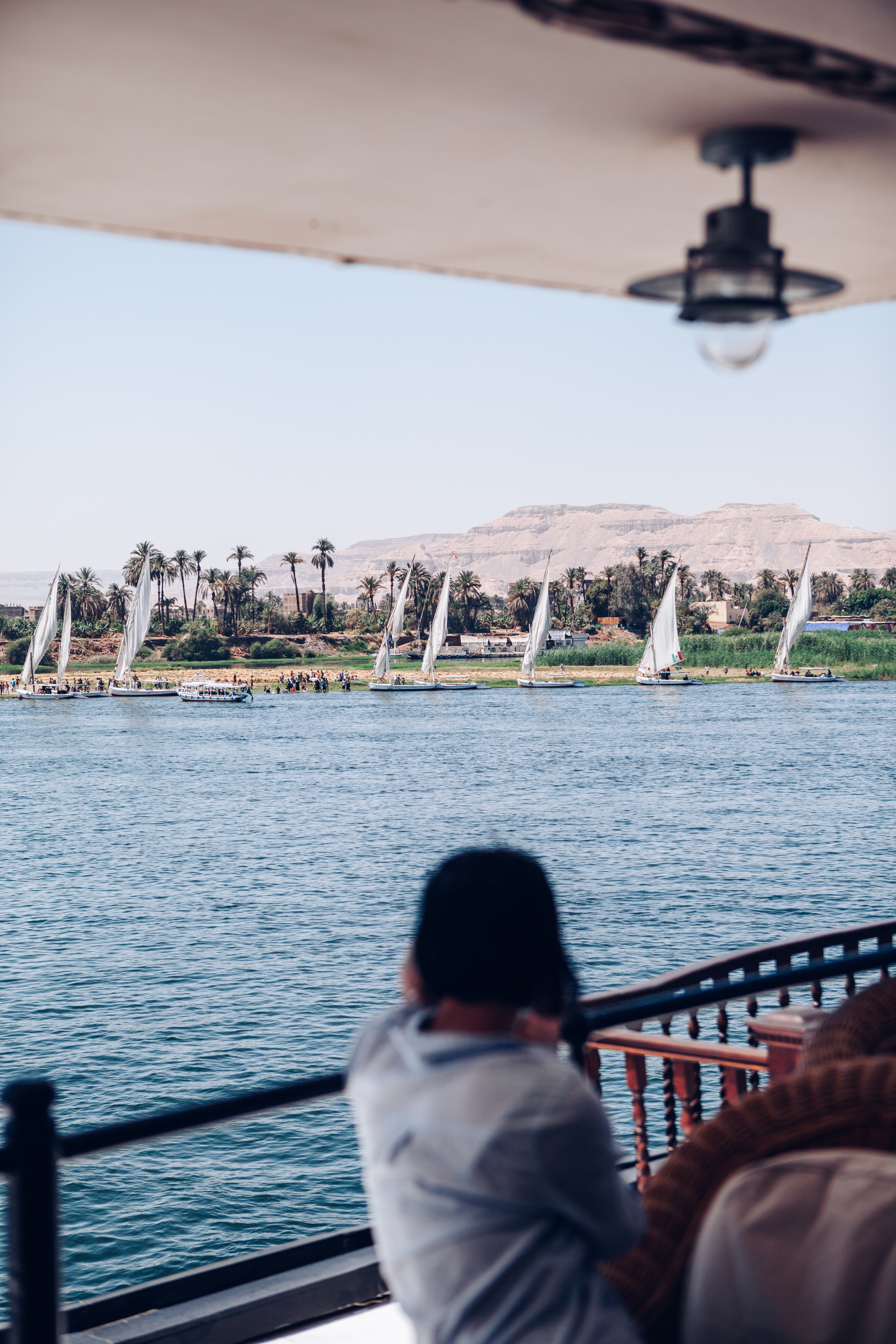 Person sits on a boat deck facing the river, with a shaded awning above and distant trees along the bank.