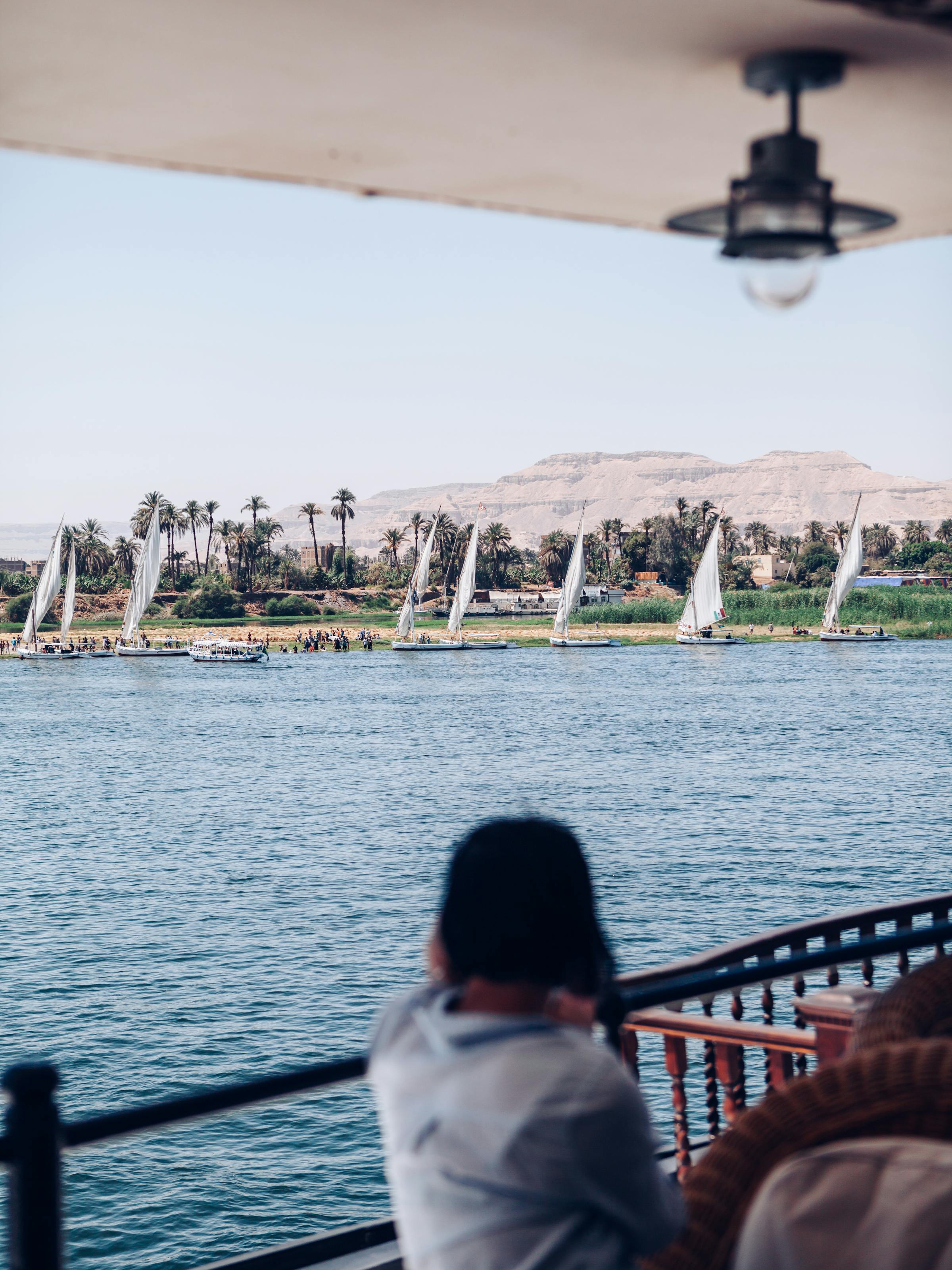 Person sits on a boat deck facing the river, with a shaded awning above and distant trees along the bank.