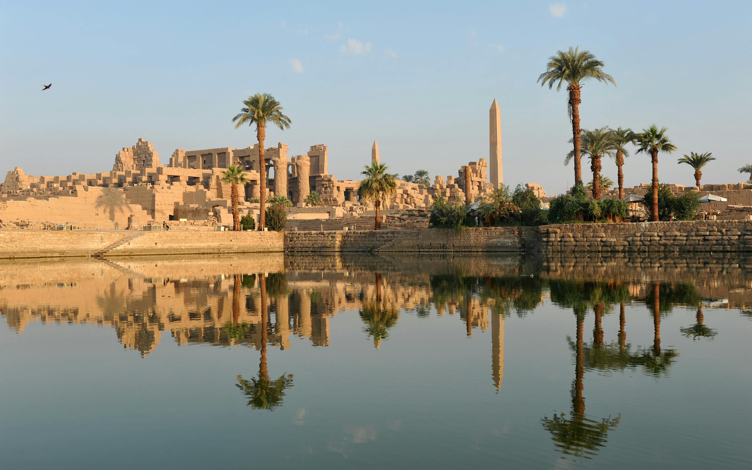 Temple ruins and tall columns reflect in still water, with palms and a bright sky mirrored on the surface.