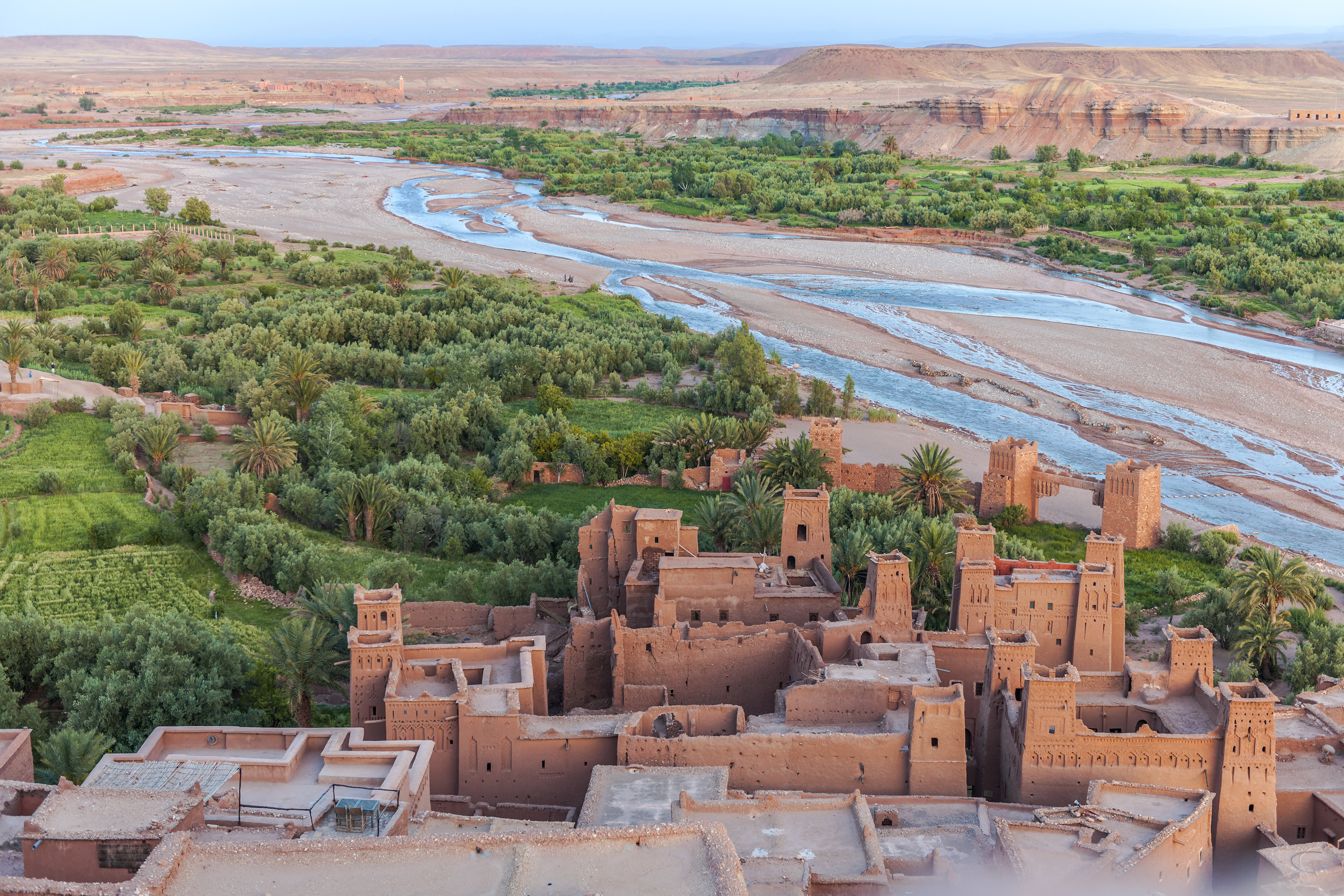High view of a mud-brick village beside a winding river and palm groves, with red cliffs and open desert beyond.