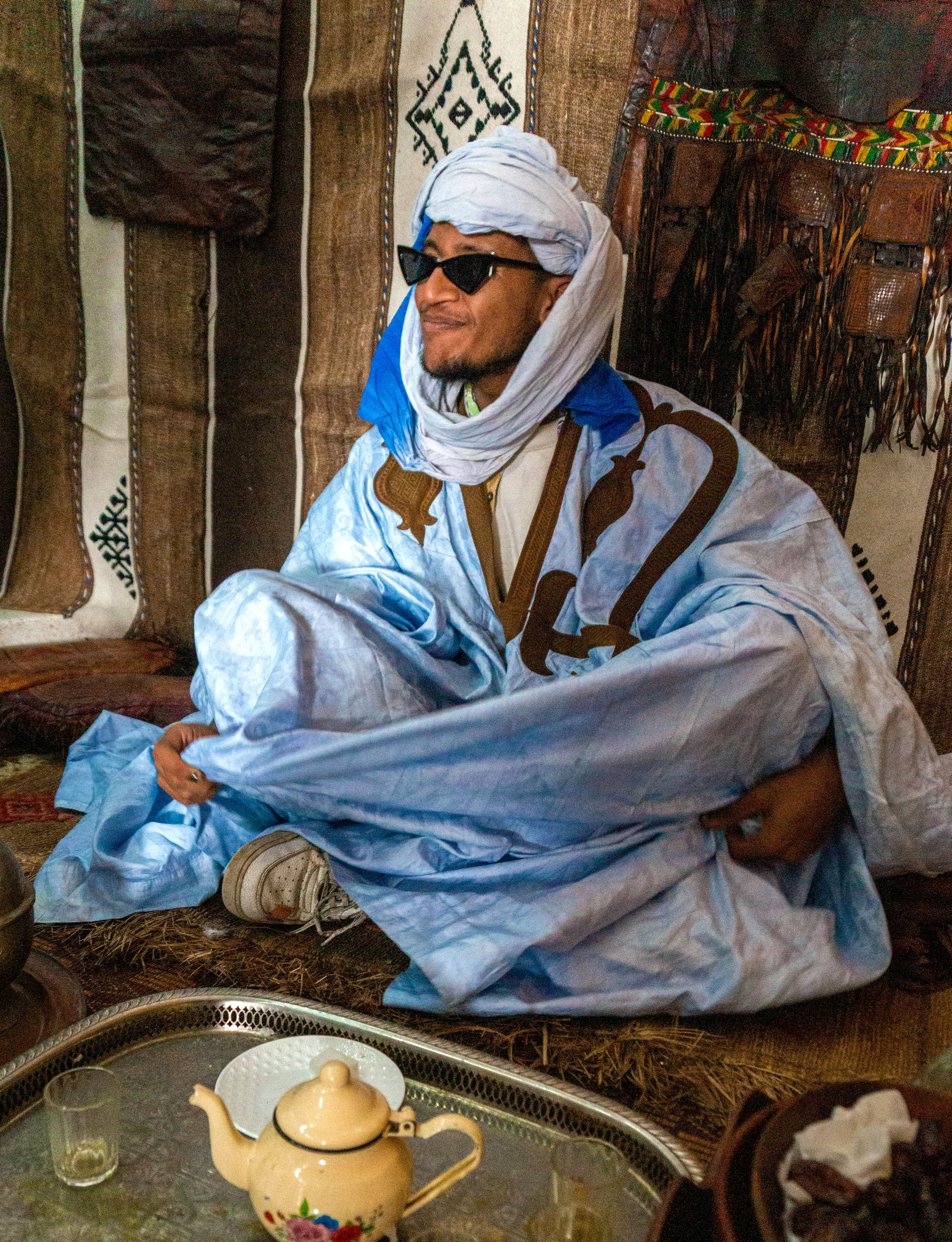 Man in a blue robe and headscarf, wearing sunglasses, sits beside a teapot and cups at an open-air café in the shade.