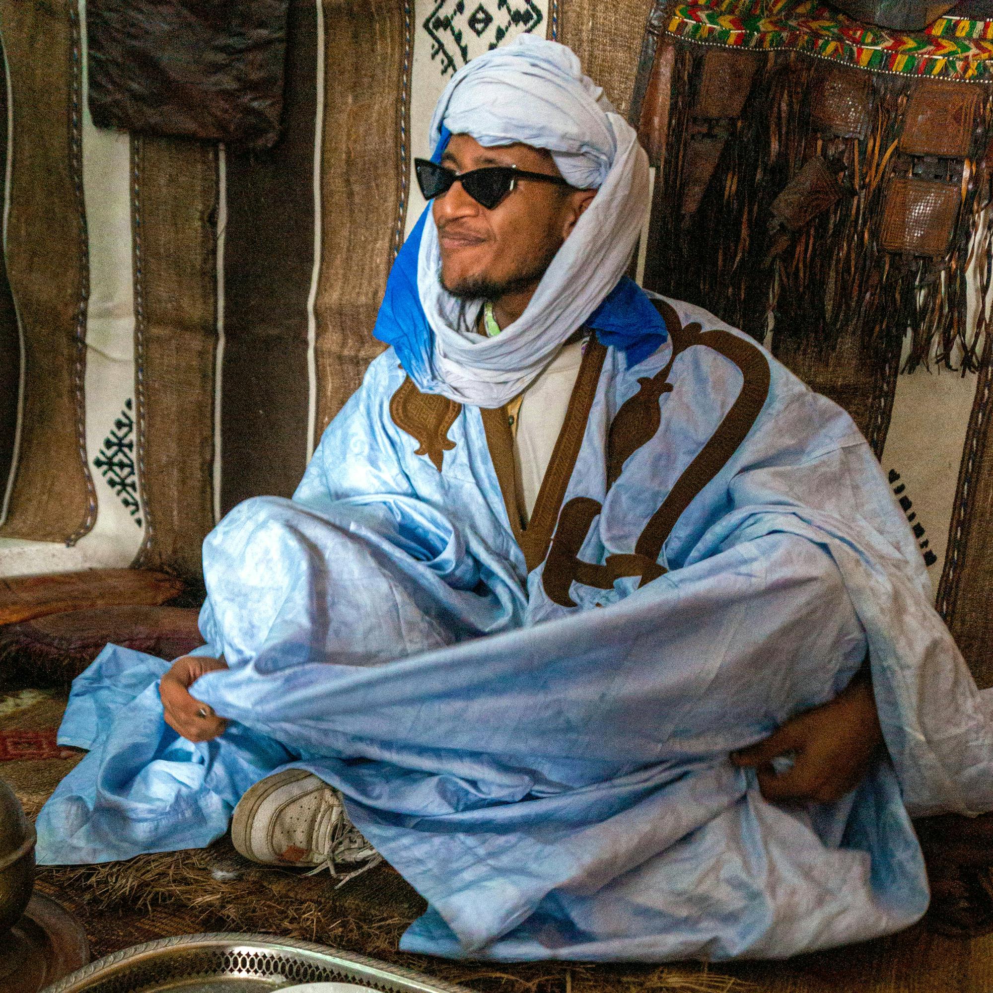 Man in a blue robe and headscarf, wearing sunglasses, sits beside a teapot and cups at an open-air café in the shade.