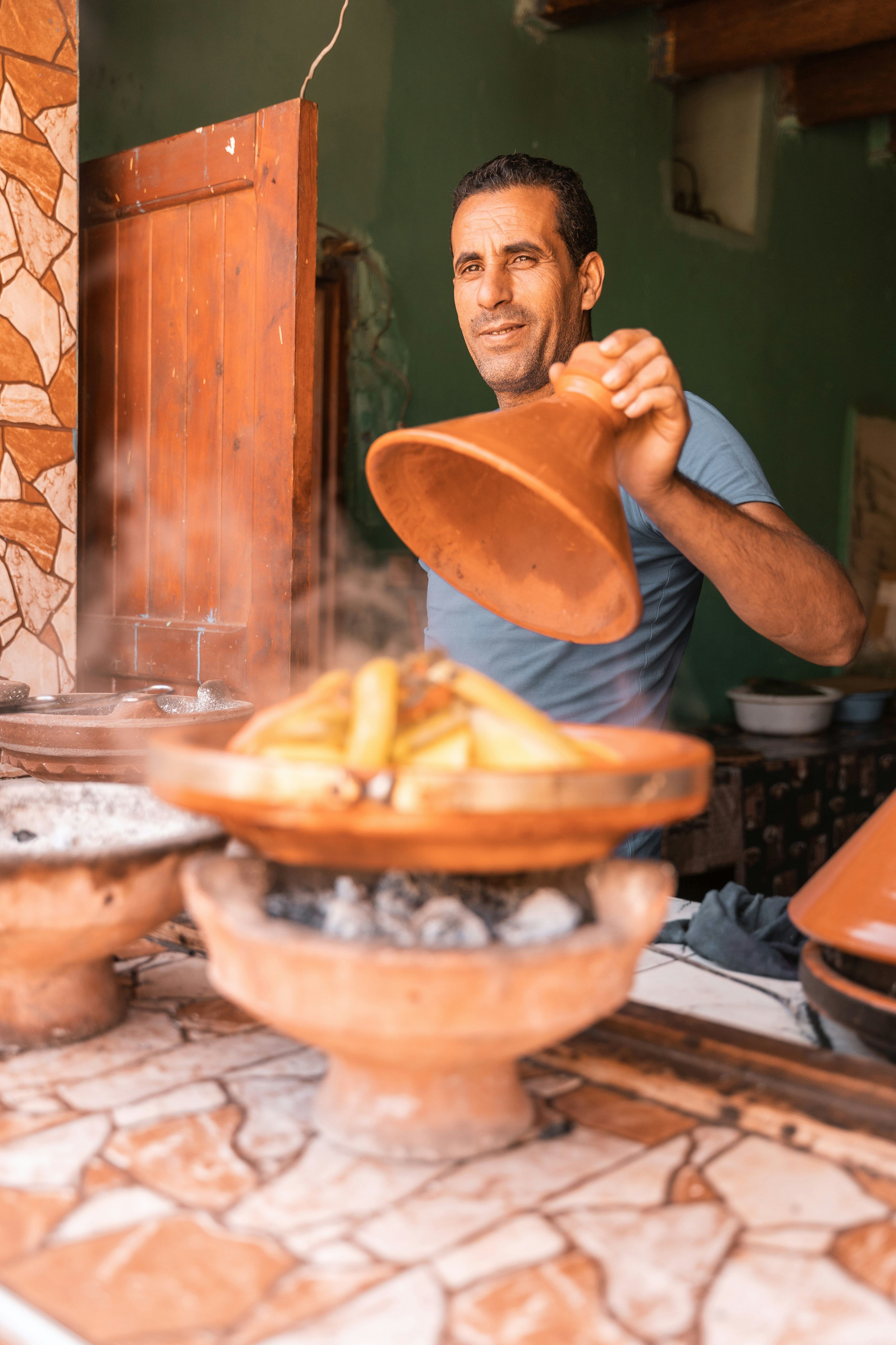 Man lifts the lid off a traditional Moroccan tagine, revealing the steaming dish beneath in warm ambient light.