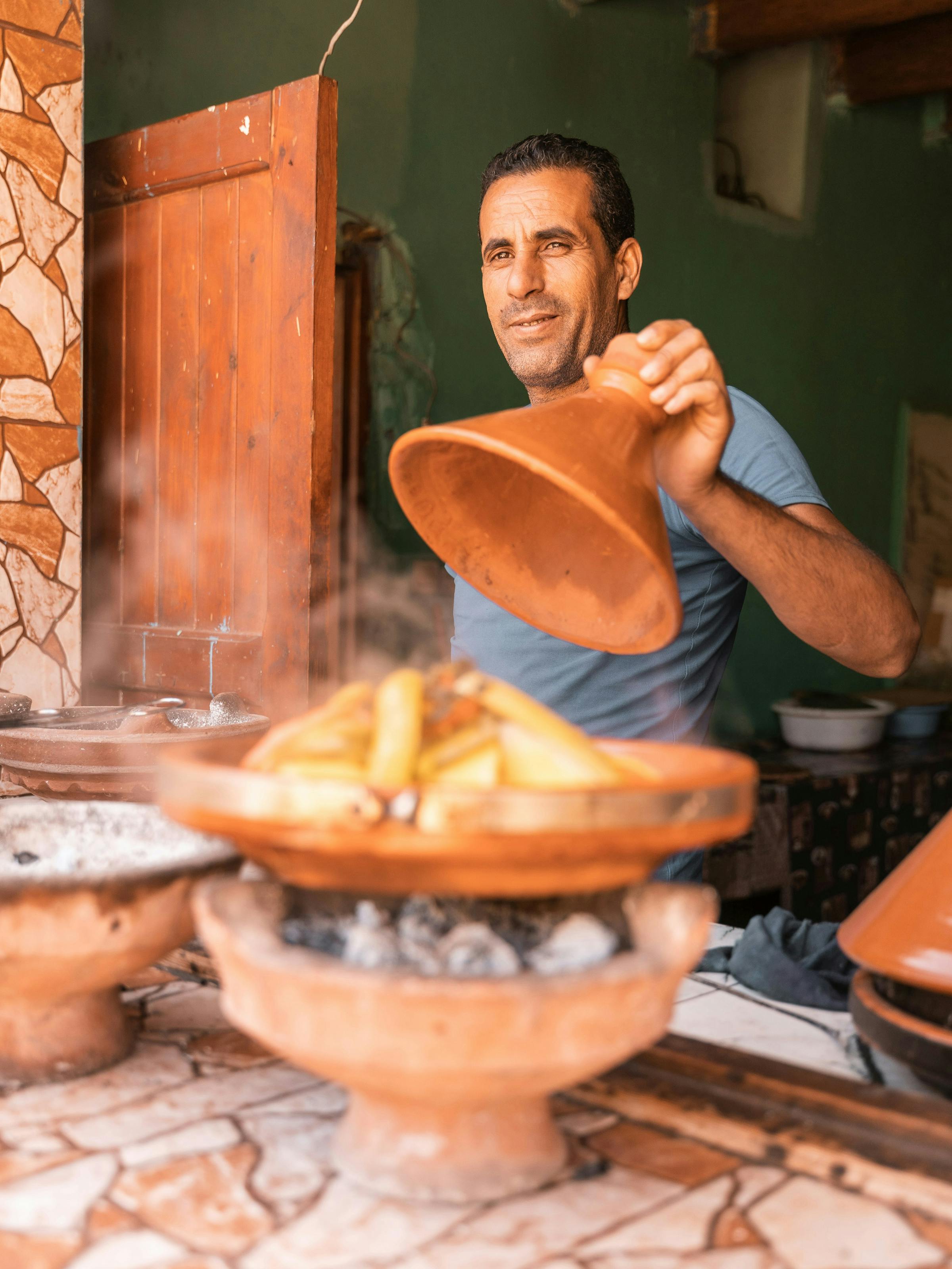Man lifts the lid off a traditional Moroccan tagine, revealing the steaming dish beneath in warm ambient light.