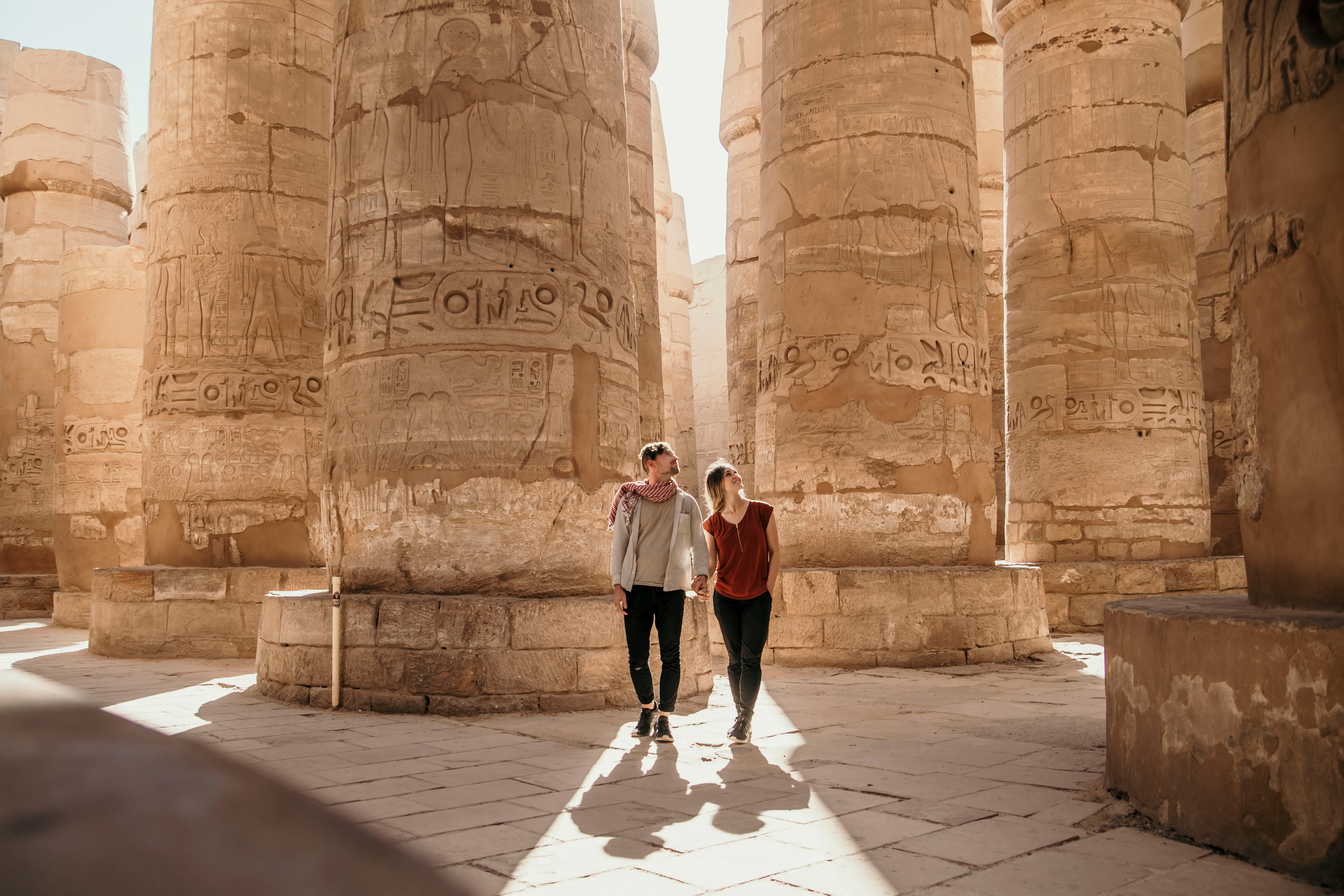 Visitors walk between massive stone columns in an ancient temple, beneath towering pillars and sharp sun shadows.