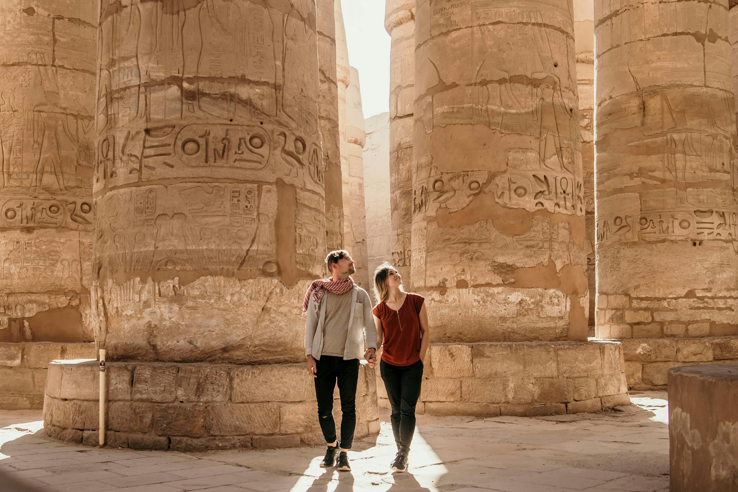 Visitors walk between massive stone columns in an ancient temple, beneath towering pillars and sharp sun shadows.
