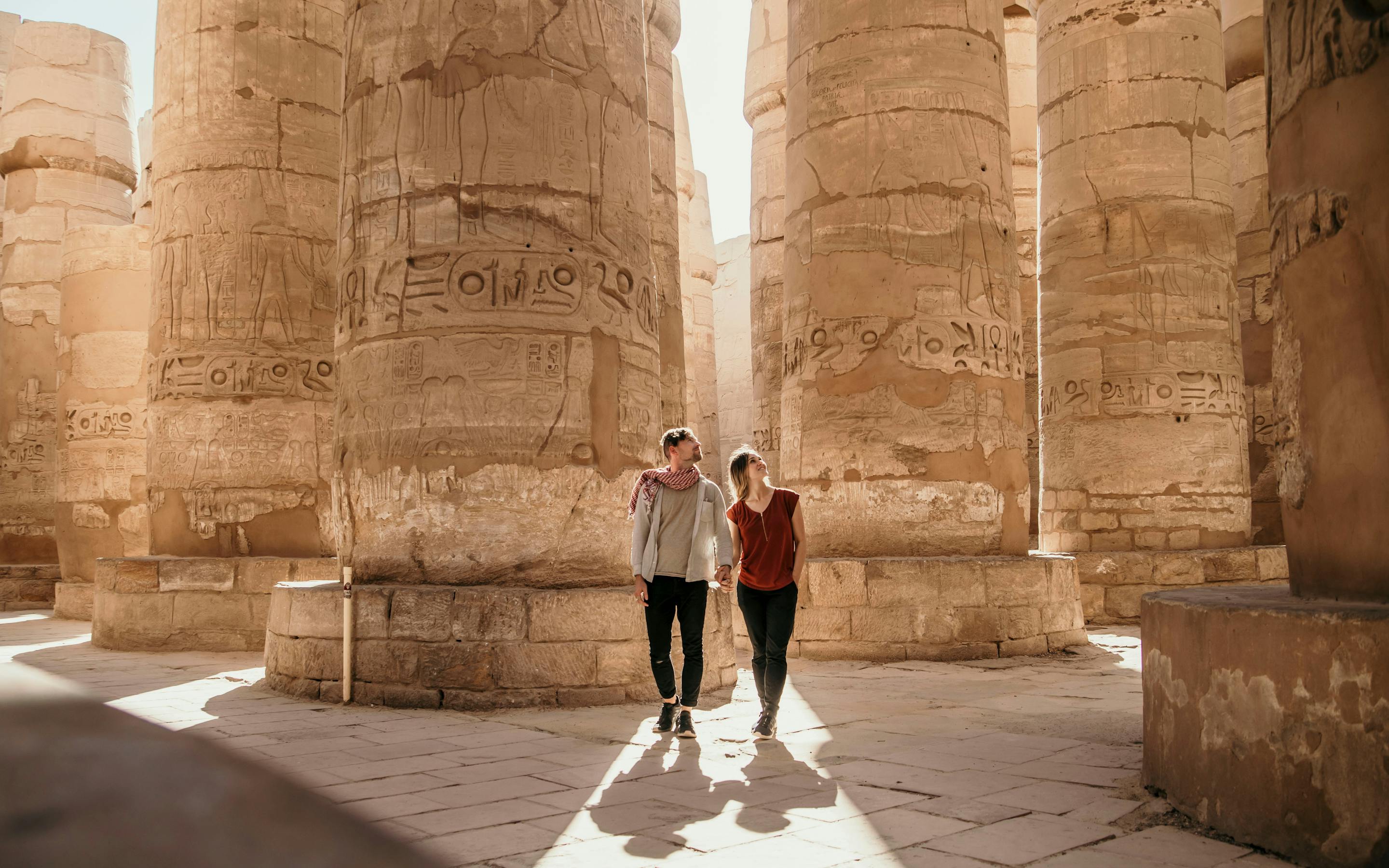 Visitors walk between massive stone columns in an ancient temple, beneath towering pillars and sharp sun shadows.
