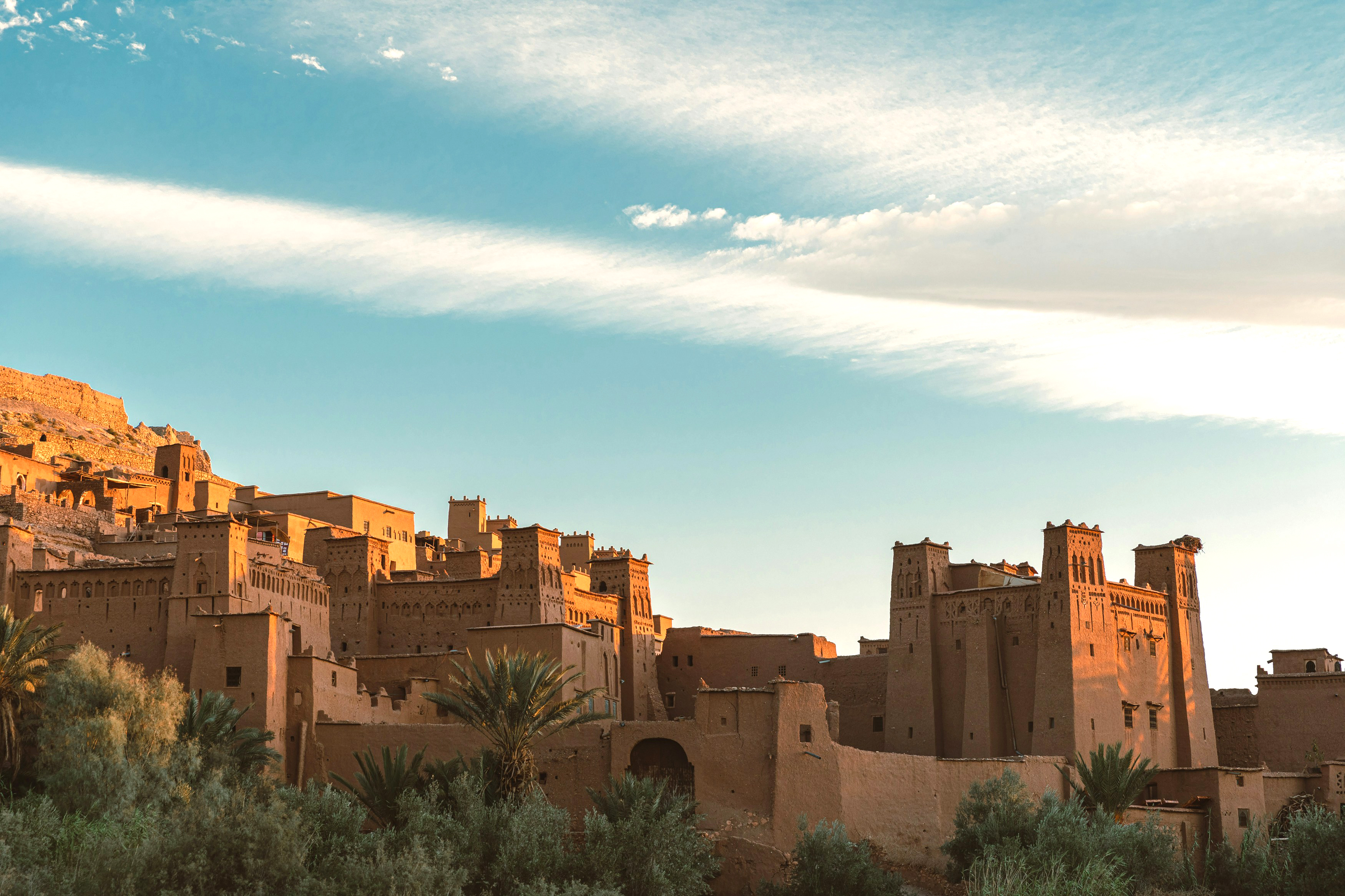 Warm sunset light falls on an earthen fortress village and palm oasis, with long clouds streaking the sky.