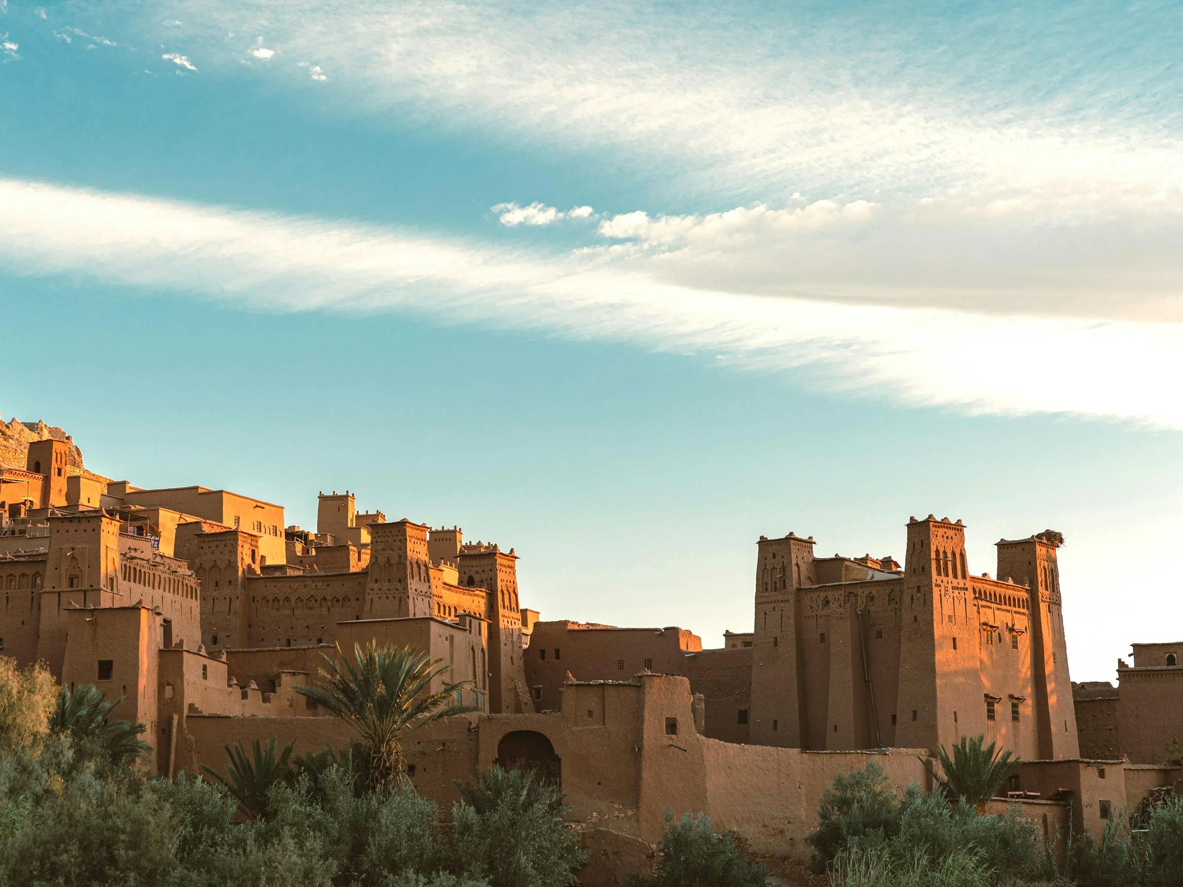 Warm sunset light falls on an earthen fortress village and palm oasis, with long clouds streaking the sky.