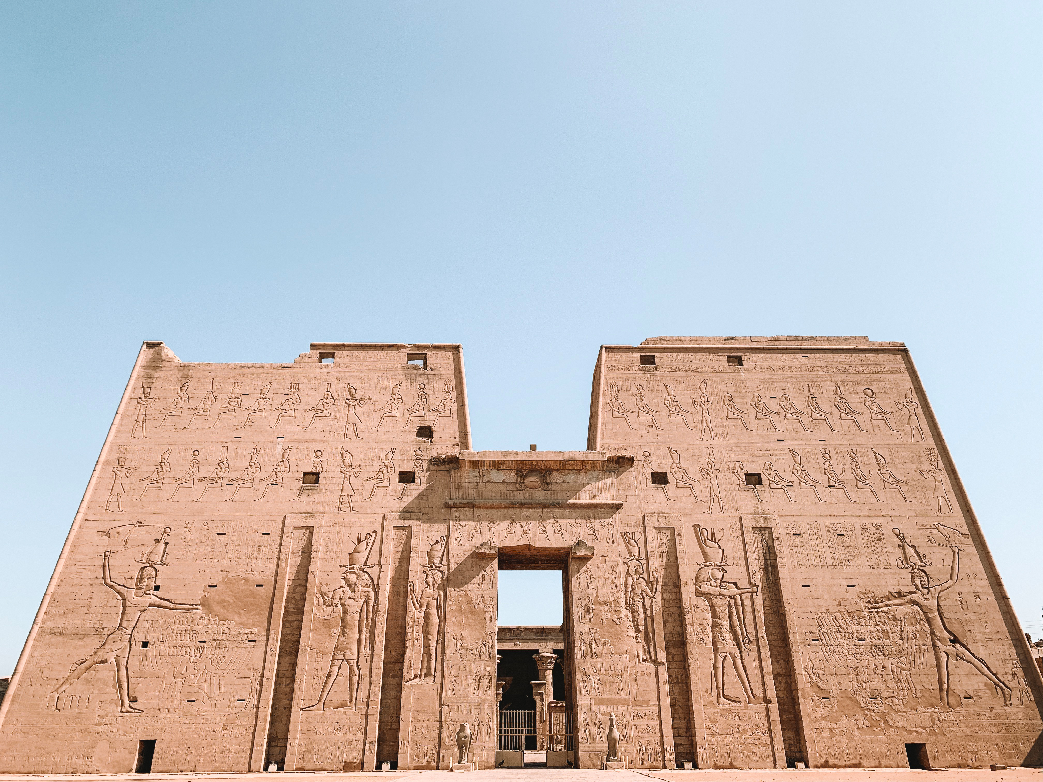 Massive stone gateway carved with hieroglyphs stands against blue sky, with a small doorway at its center.