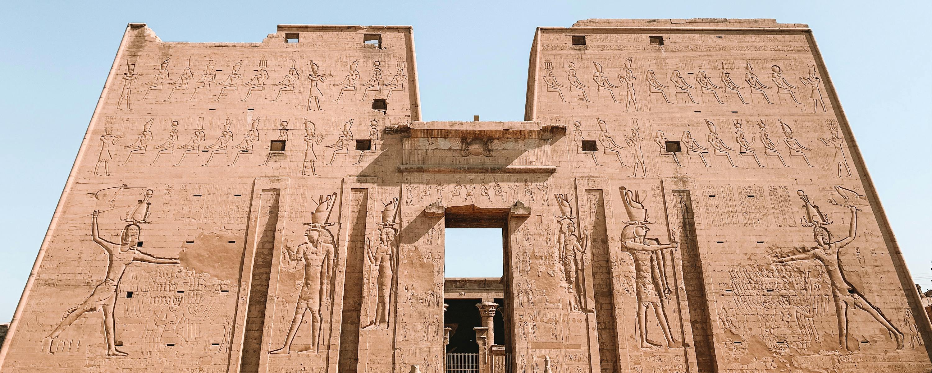 Massive stone gateway carved with hieroglyphs stands against blue sky, with a small doorway at its center.