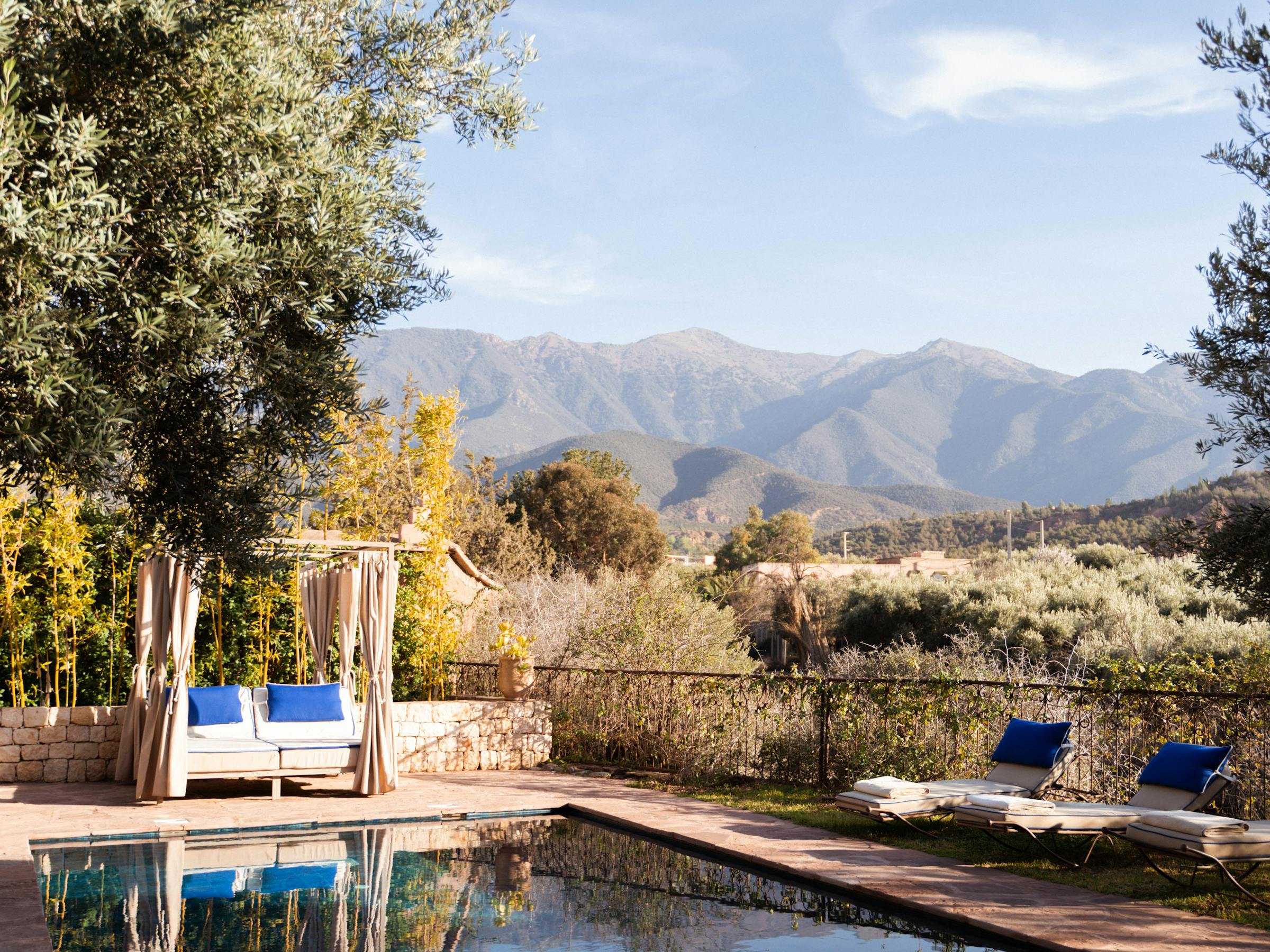 Rectangular pool reflects trees and mountains, with lounge chairs and umbrellas arranged along a stone deck.