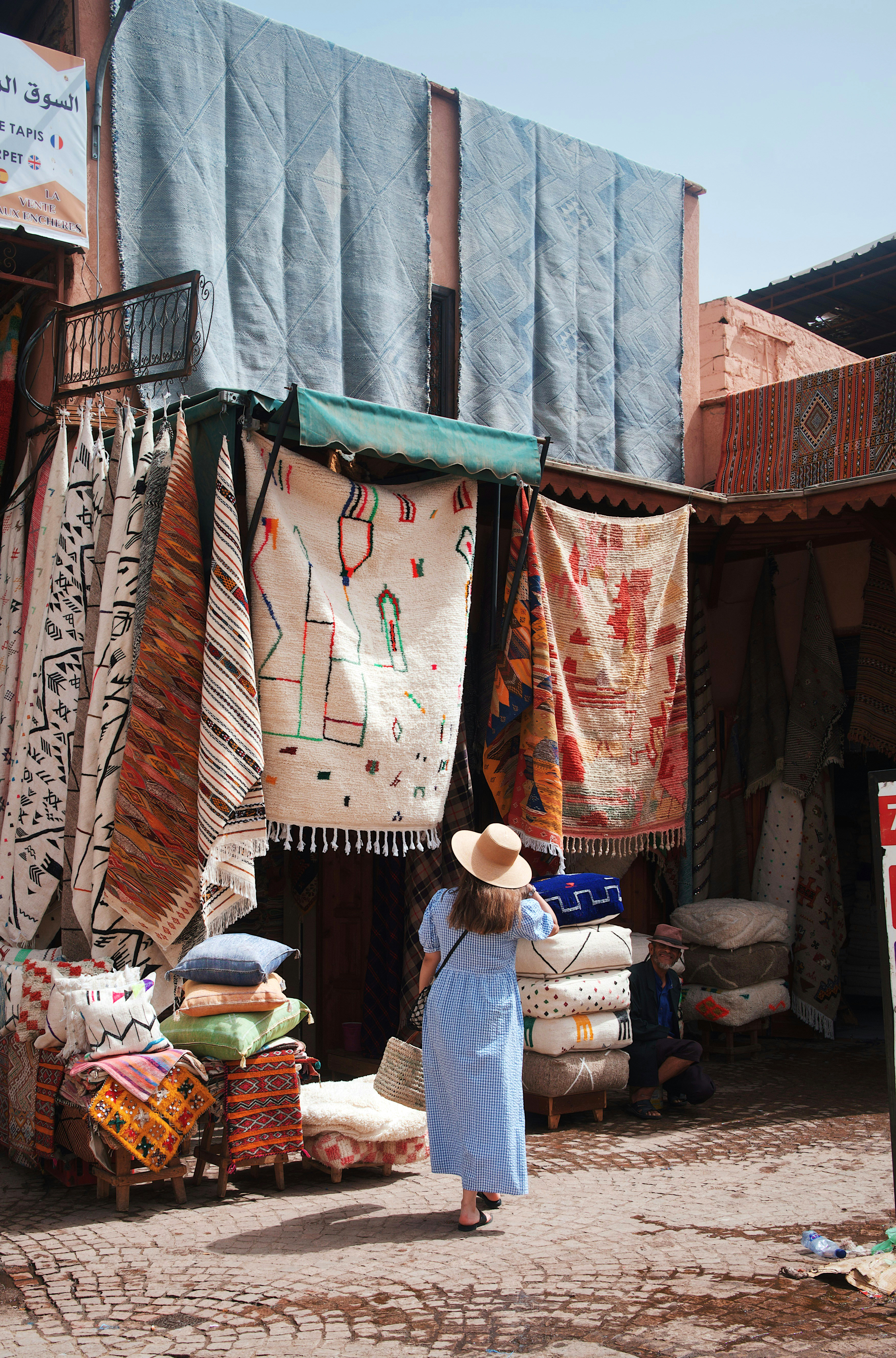 Street market scene with hanging rugs and woven textiles, as a person in a white robe stands near the stall entrance.