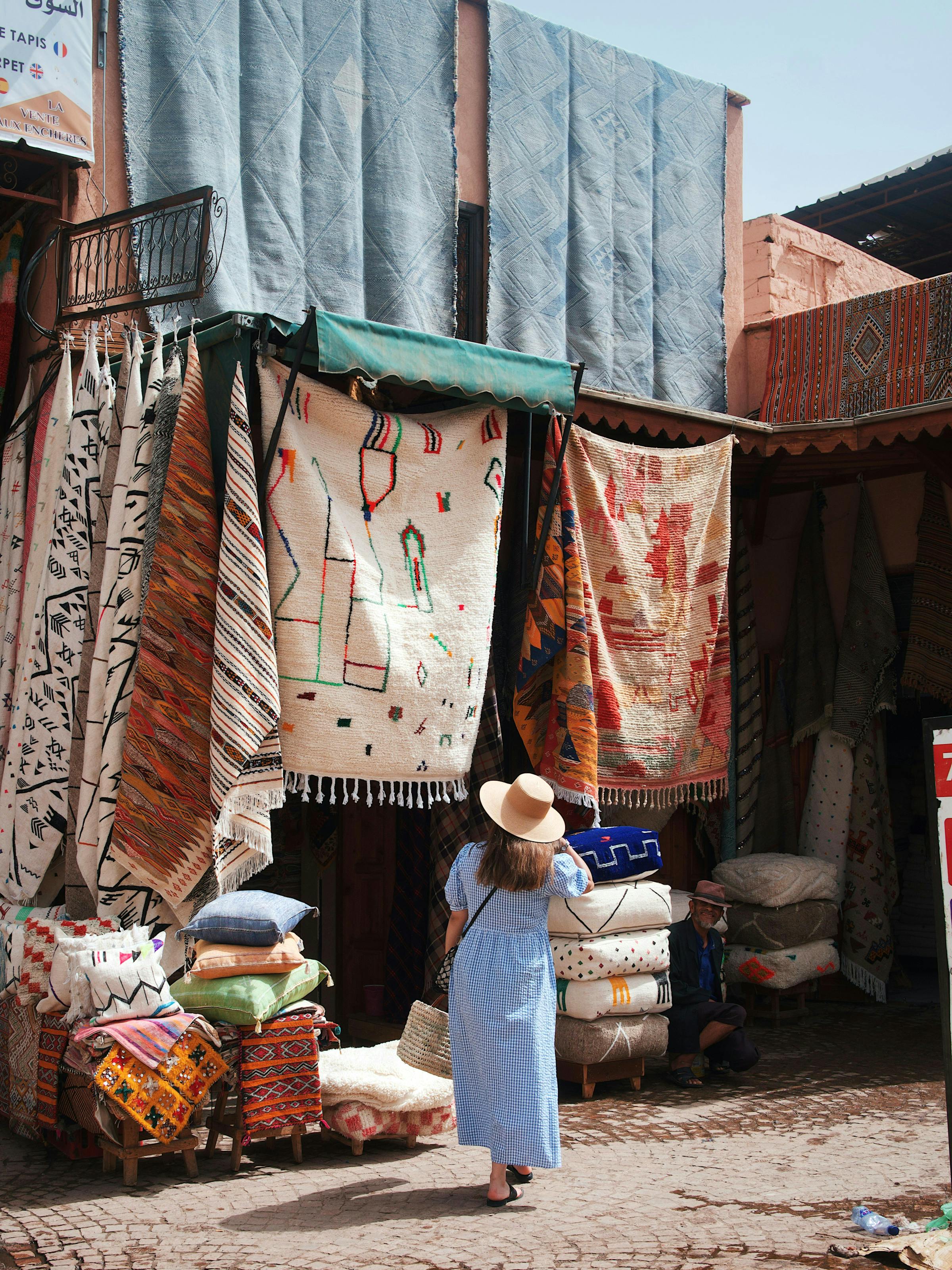 Street market scene with hanging rugs and woven textiles, as a person in a white robe stands near the stall entrance.