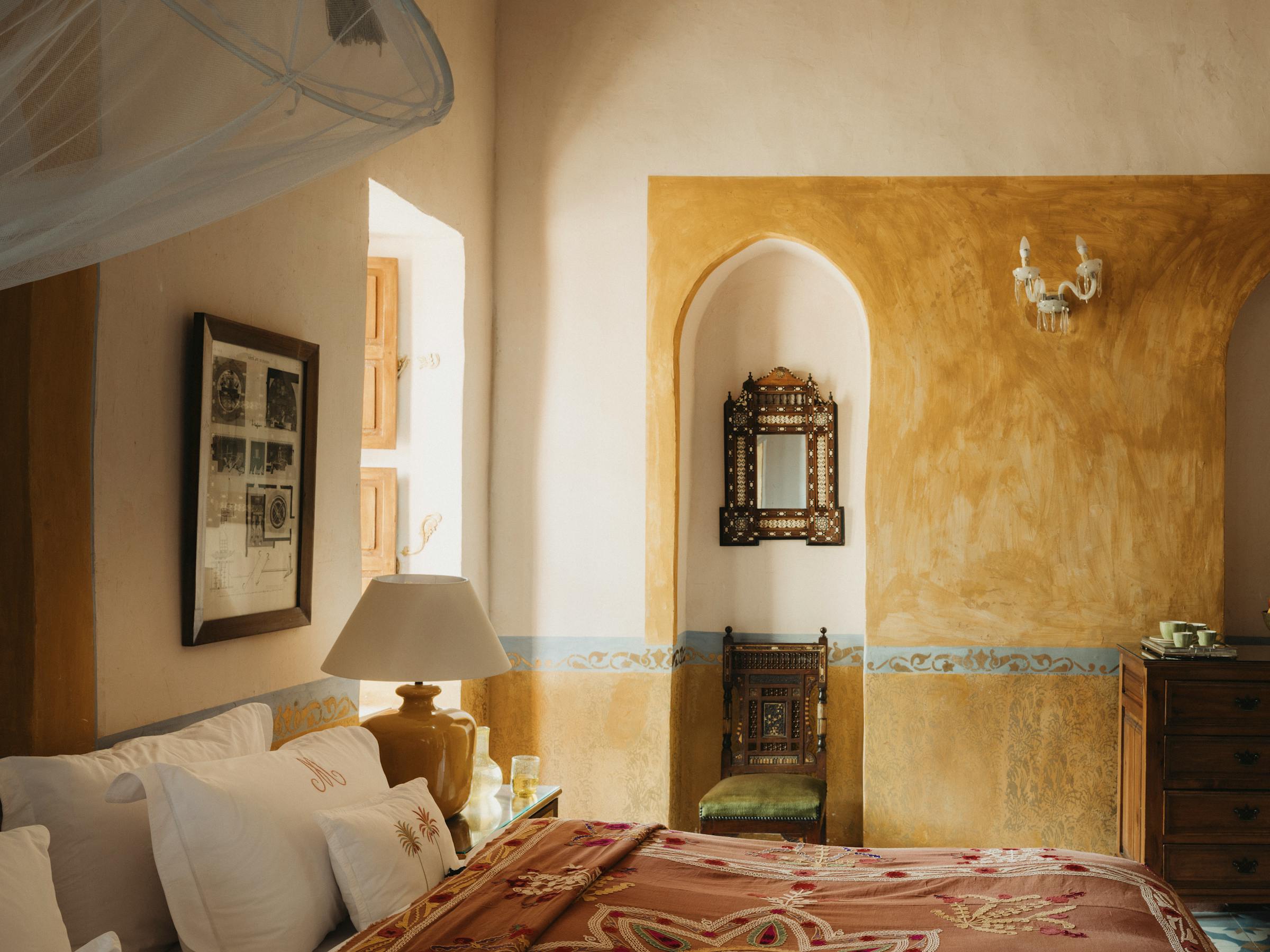 Cozy hotel room with a patterned bedspread, arched doorways, tiled floors, and warm plaster walls lit by daylight.