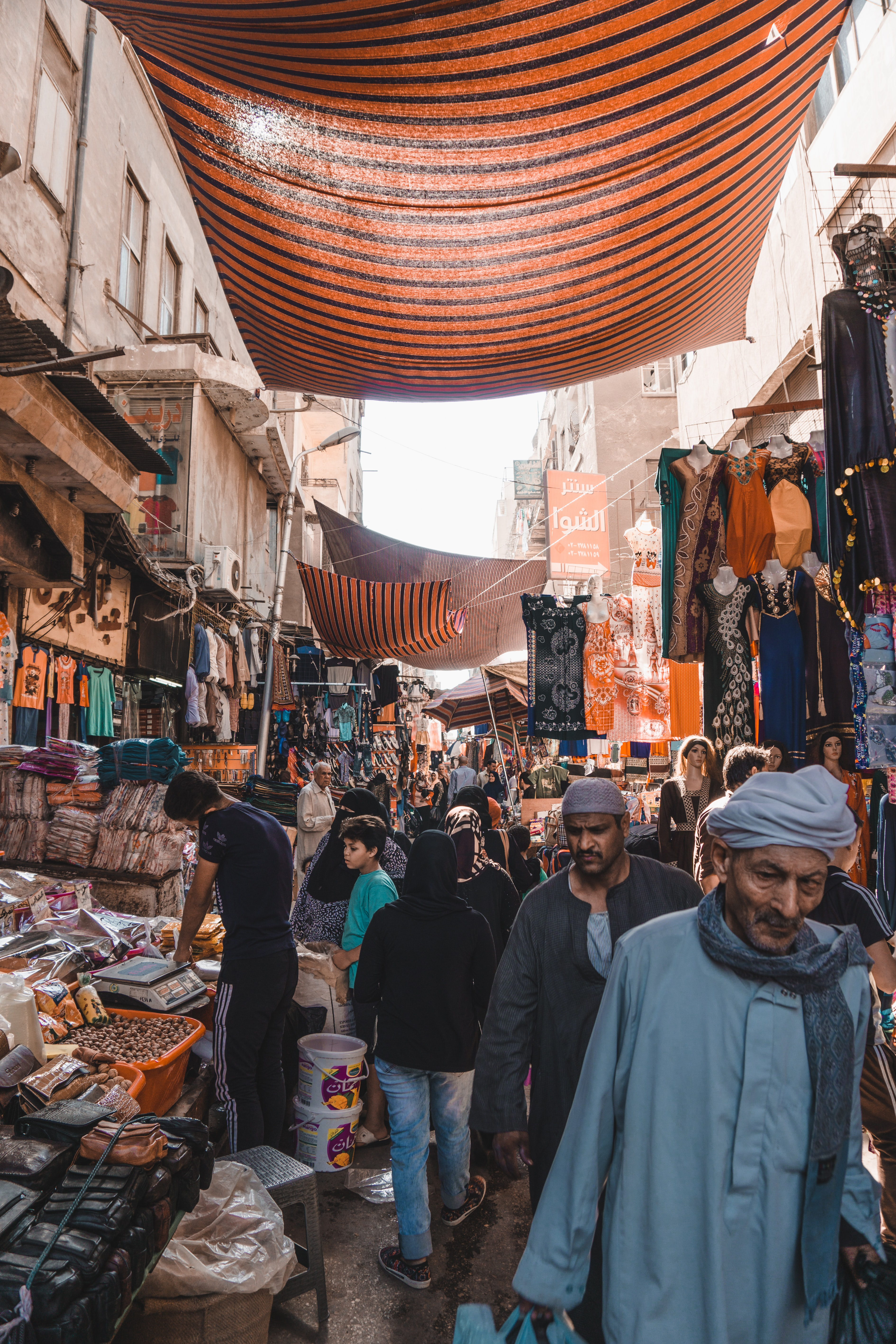 Busy market street under striped fabric canopies, with shoppers walking past stalls as daylight filters through.