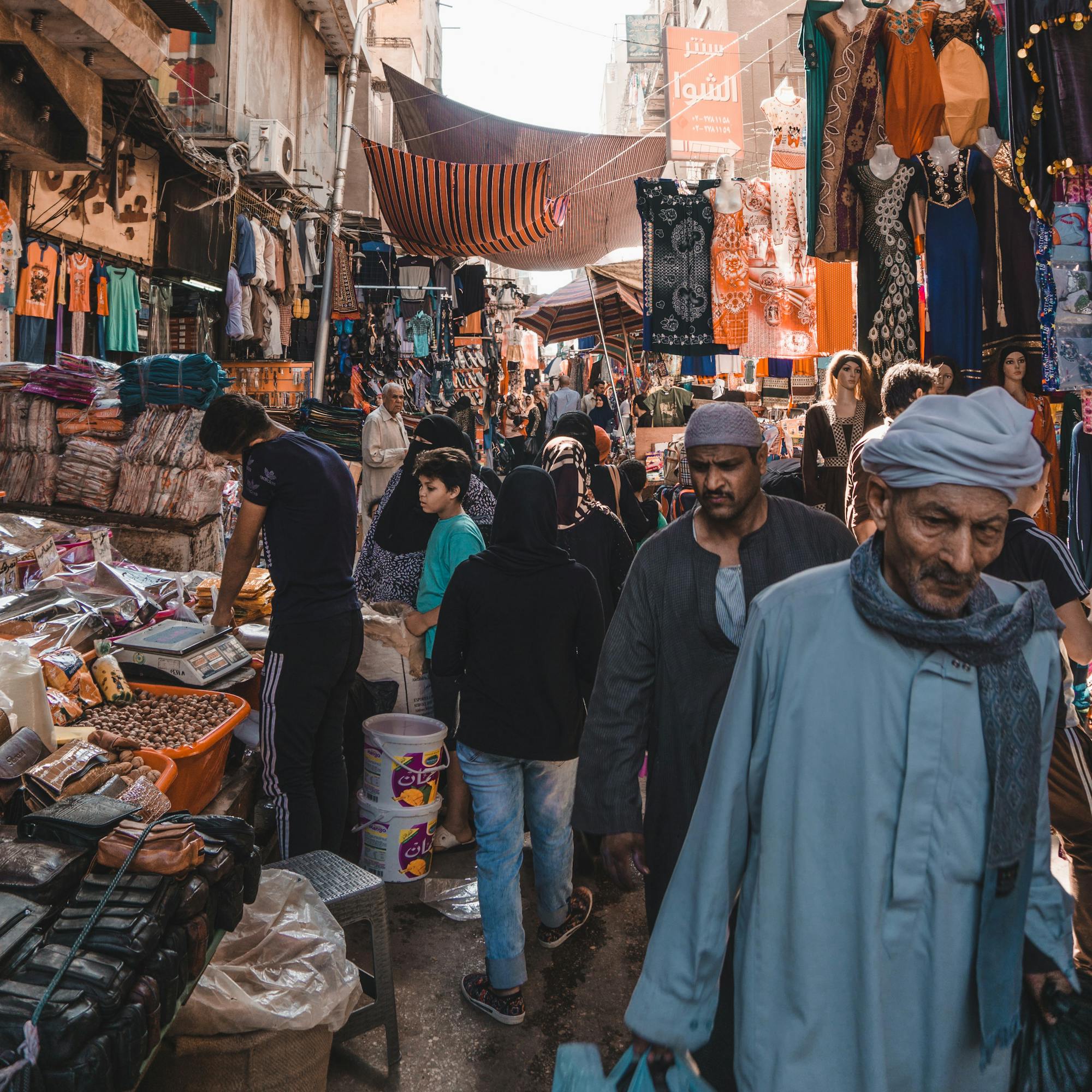 Busy market street under striped fabric canopies, with shoppers walking past stalls as daylight filters through.