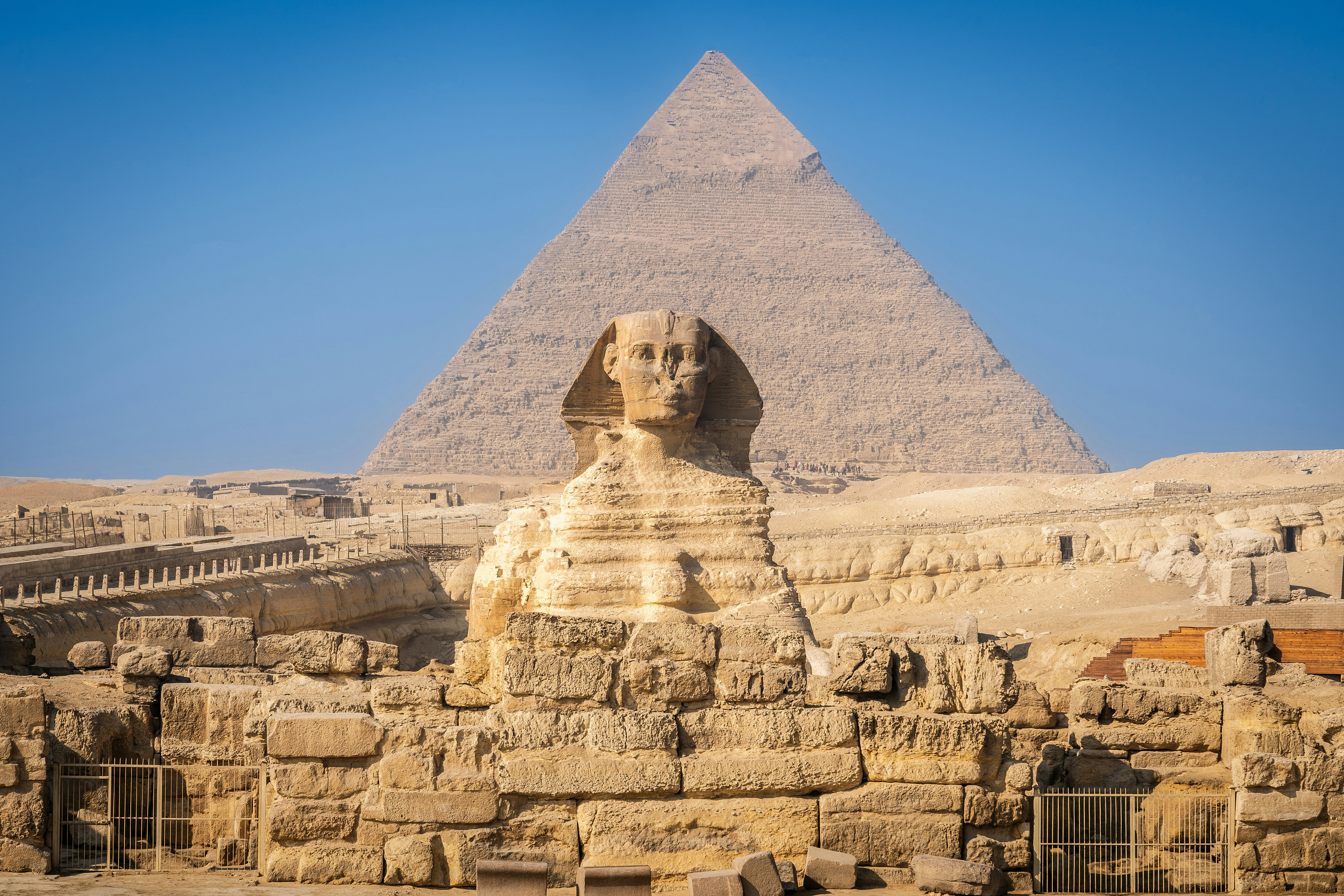 The Great Sphinx sits in front of a pyramid, with stepped stone terraces in bright sun under a deep blue sky.