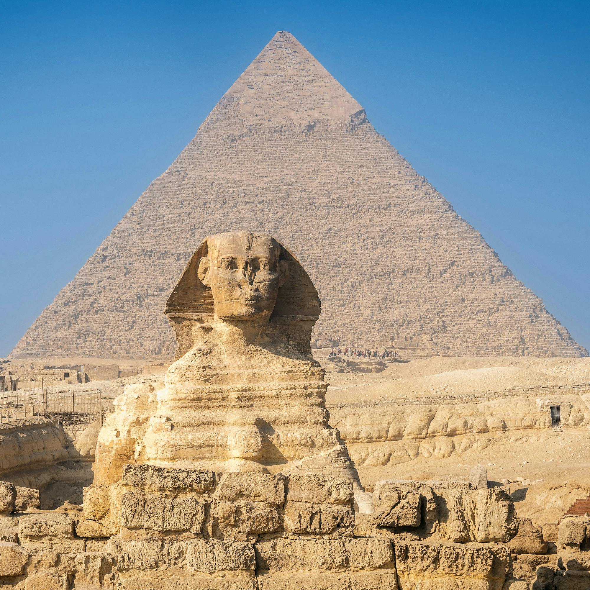The Great Sphinx sits in front of a pyramid, with stepped stone terraces in bright sun under a deep blue sky.