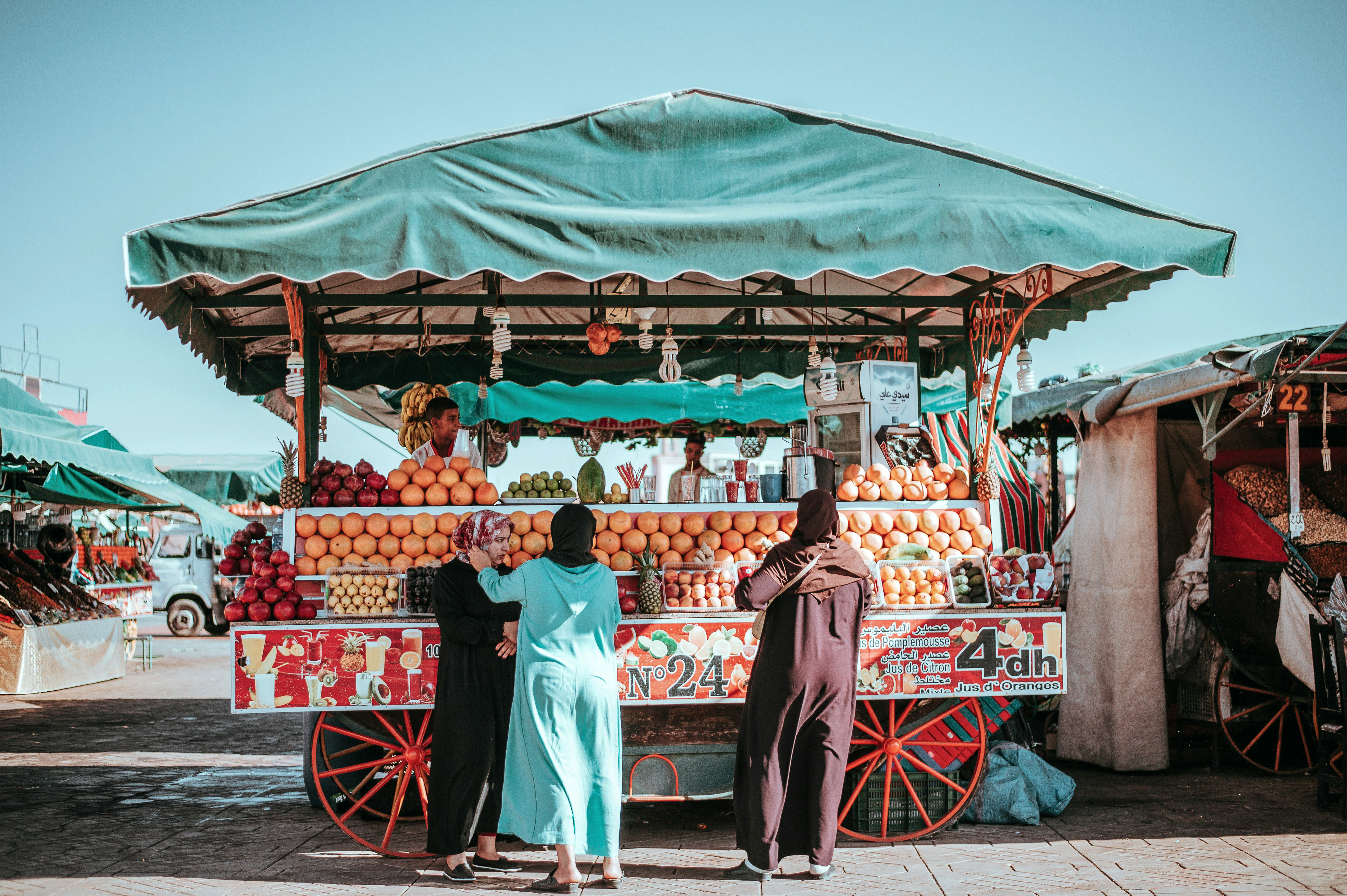 Open-air fruit cart under a green canopy, with vendors standing beside stacked produce and bright street signs.