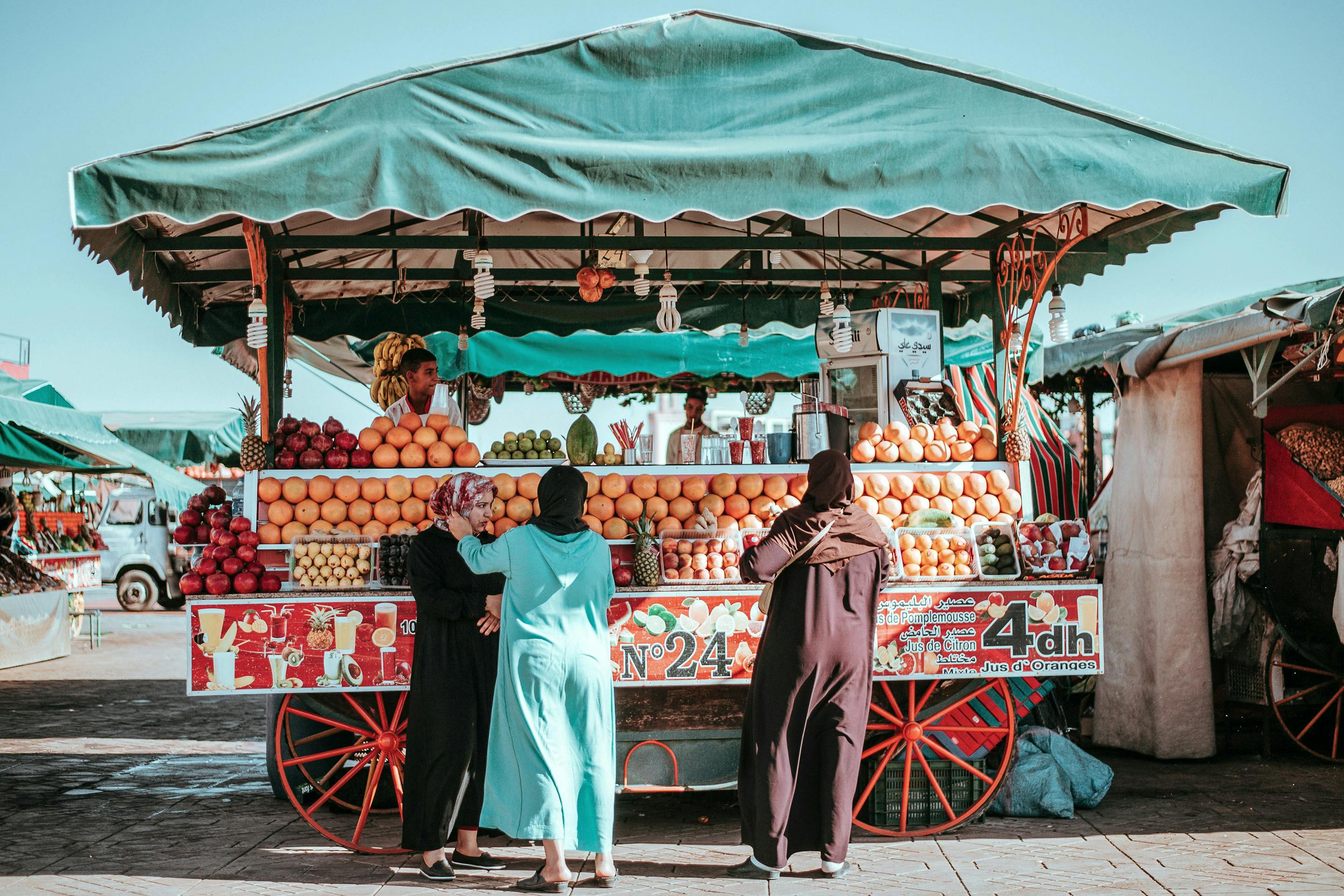 Open-air fruit cart under a green canopy, with vendors standing beside stacked produce and bright street signs.
