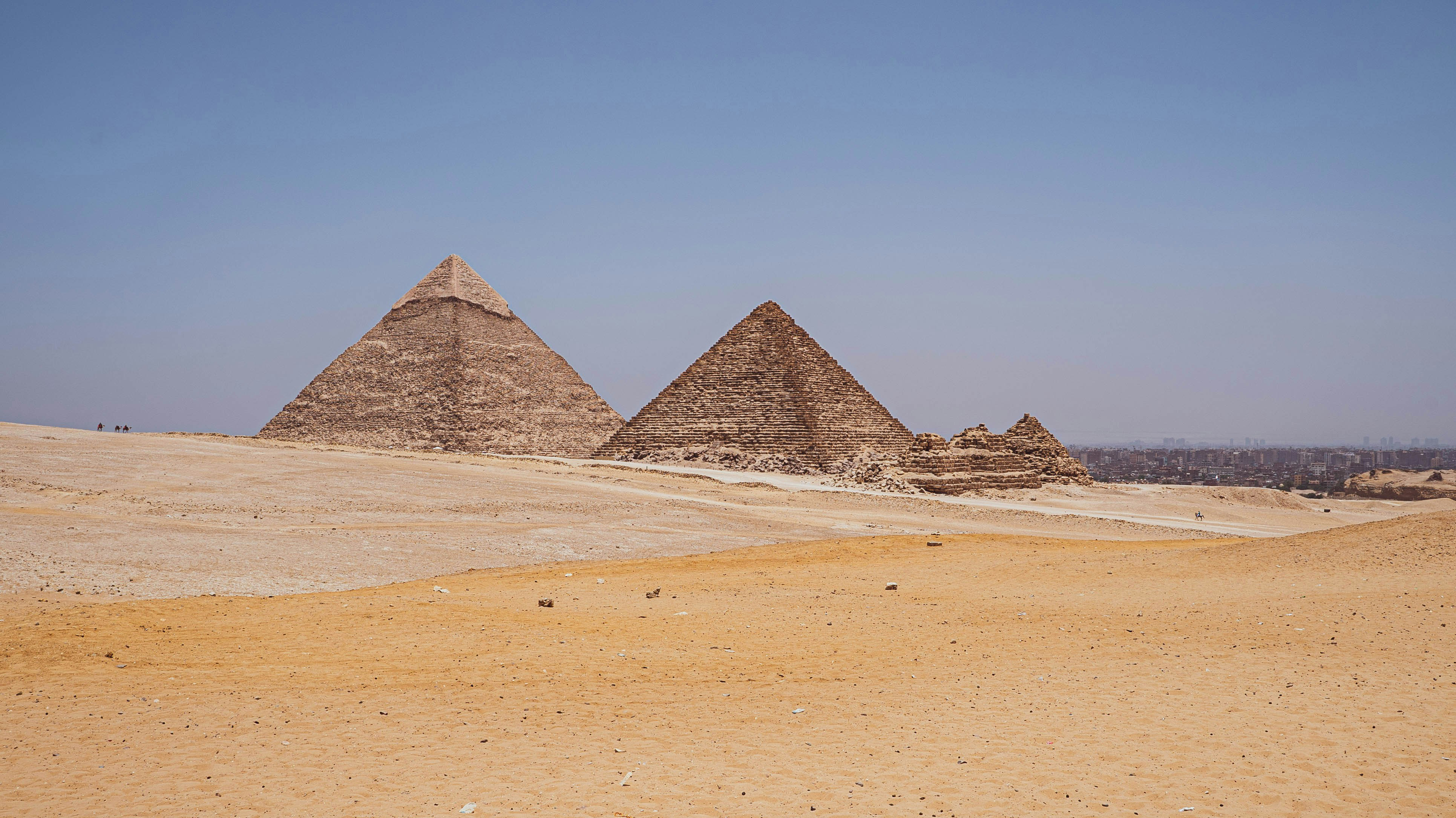 Three pyramids rise from open desert sands, with wind-swept dunes in the foreground and clear blue sky above.