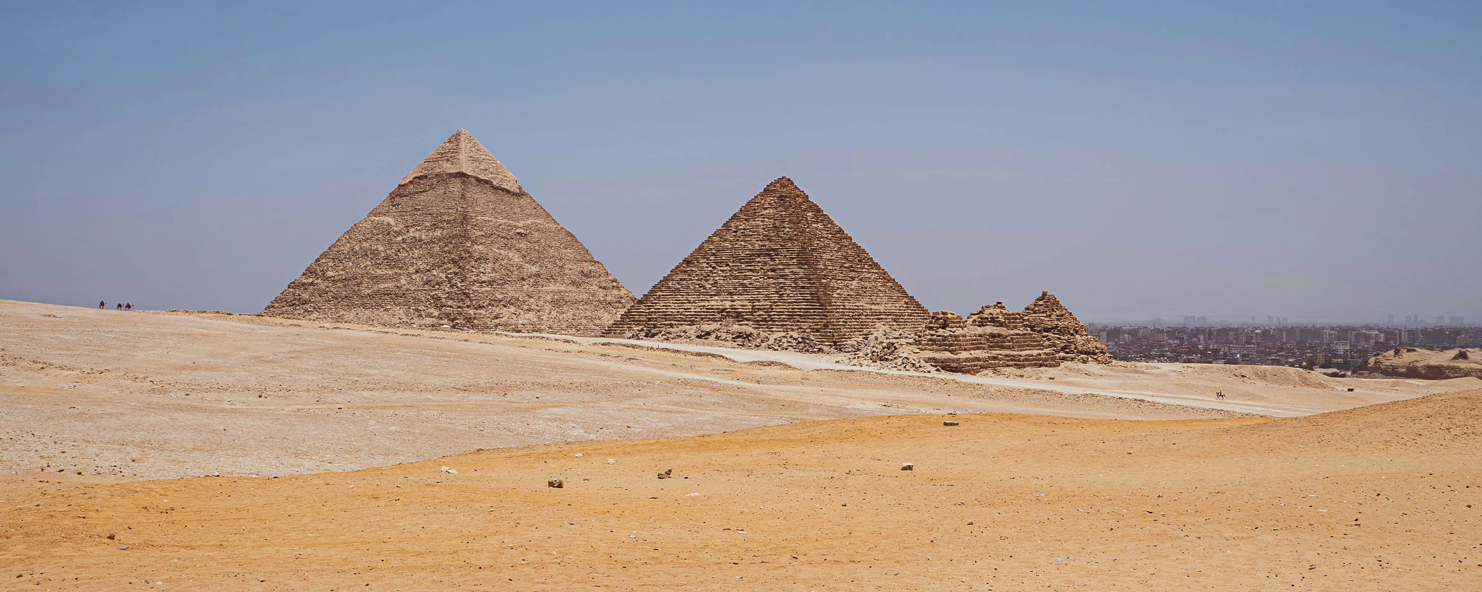 Three pyramids rise from open desert sands, with wind-swept dunes in the foreground and clear blue sky above.