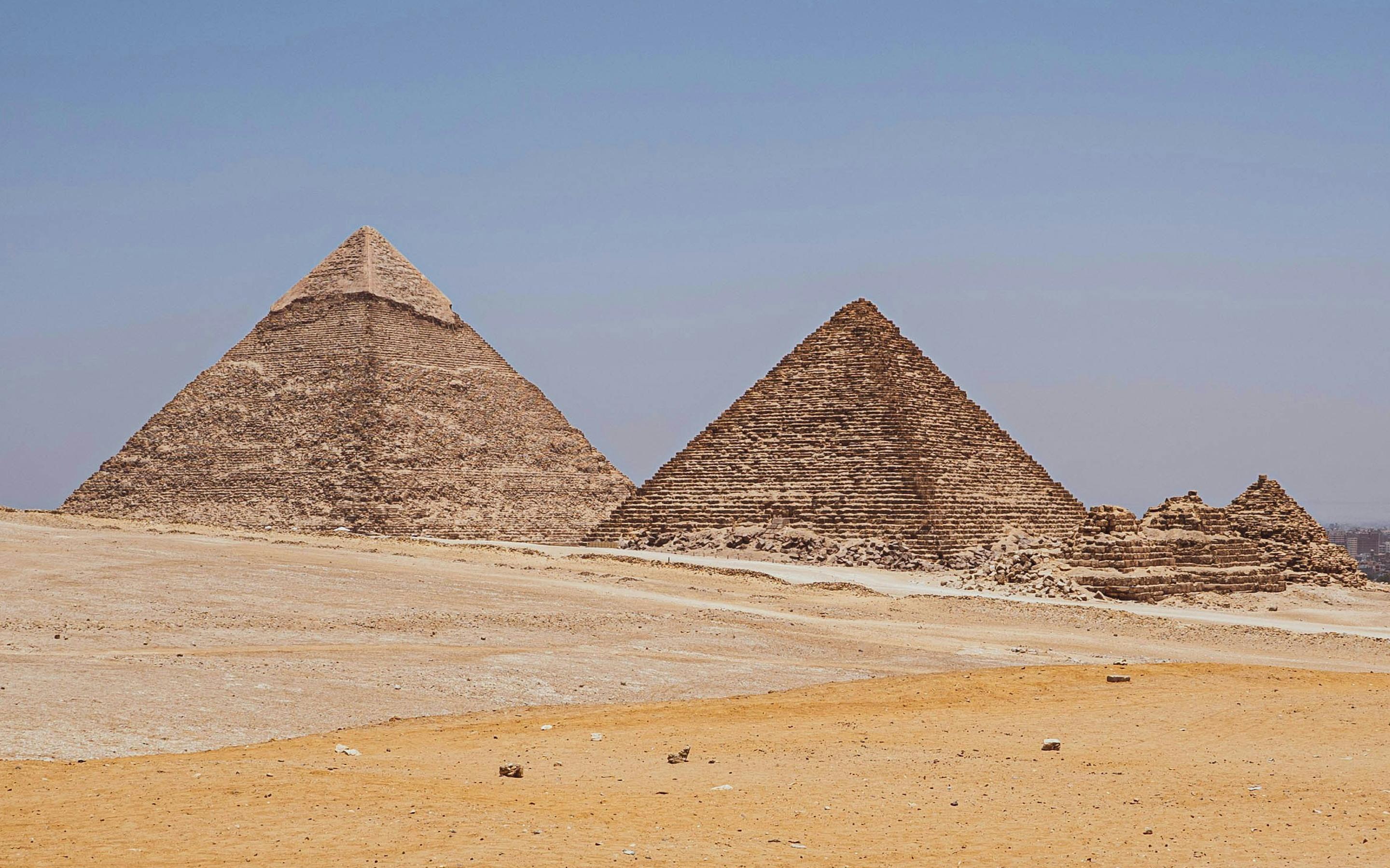 Three pyramids rise from open desert sands, with wind-swept dunes in the foreground and clear blue sky above.