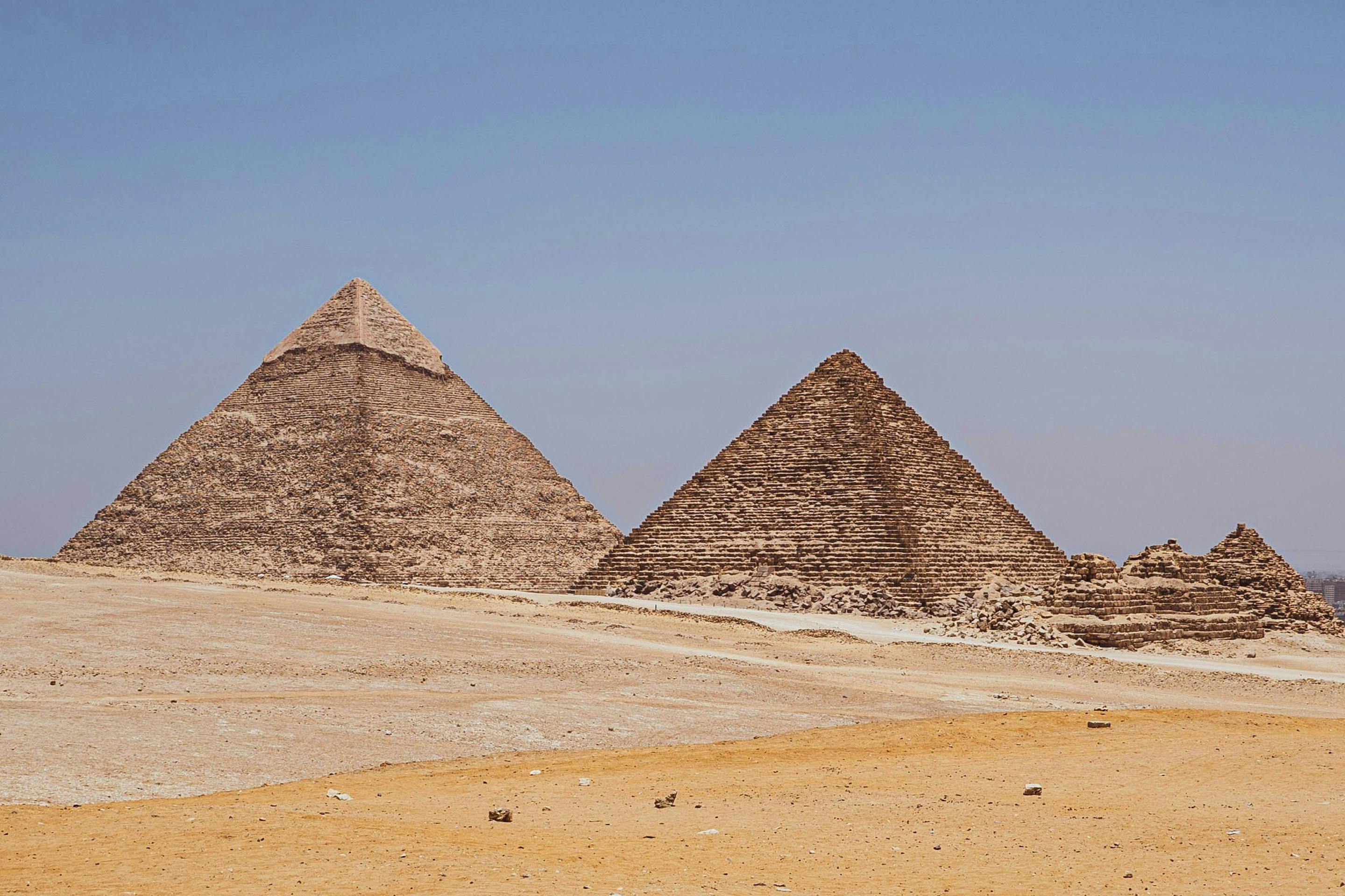 Three pyramids rise from open desert sands, with wind-swept dunes in the foreground and clear blue sky above.