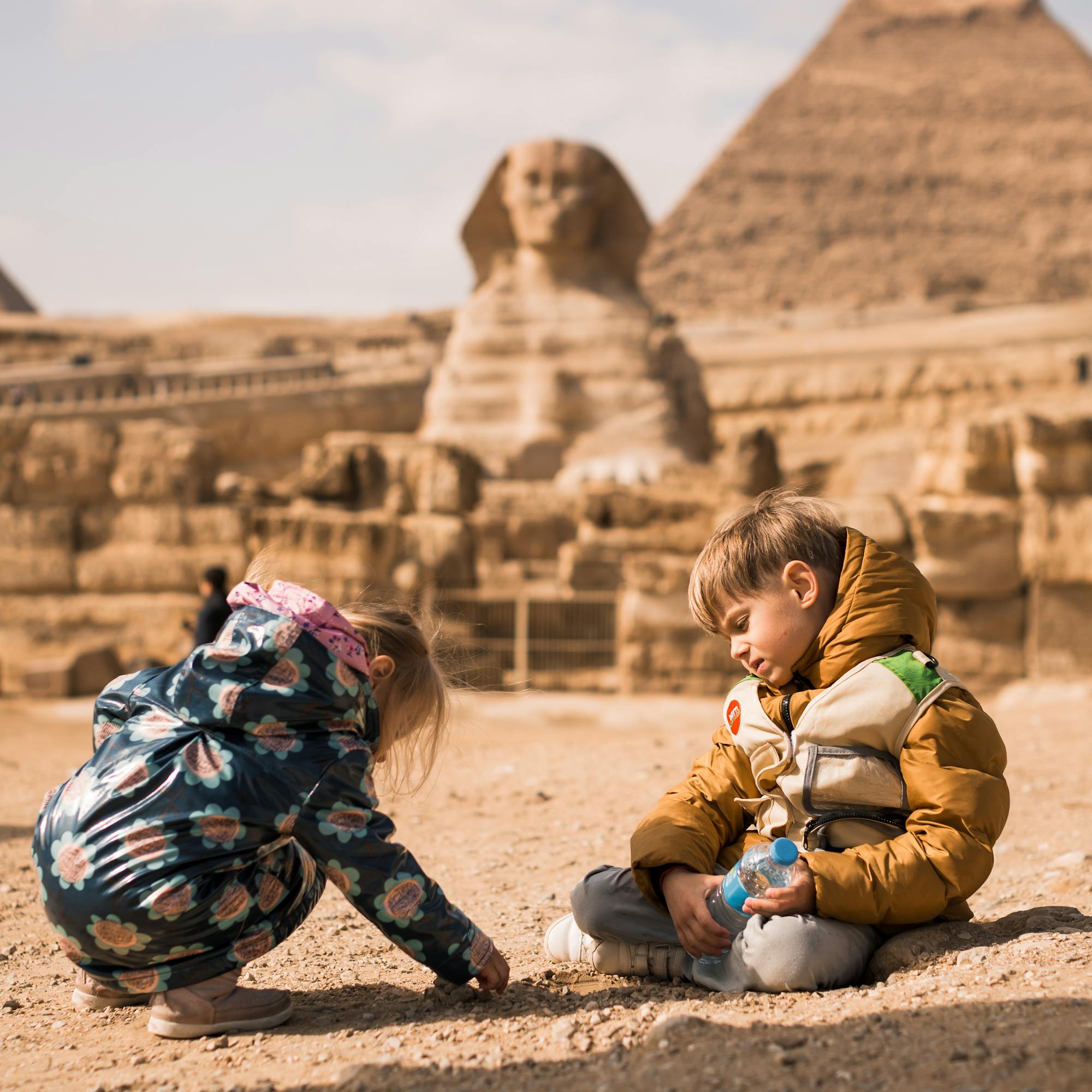 Tourists sit on sandy ground near the Great Sphinx and a pyramid, with camels and desert haze behind them.
