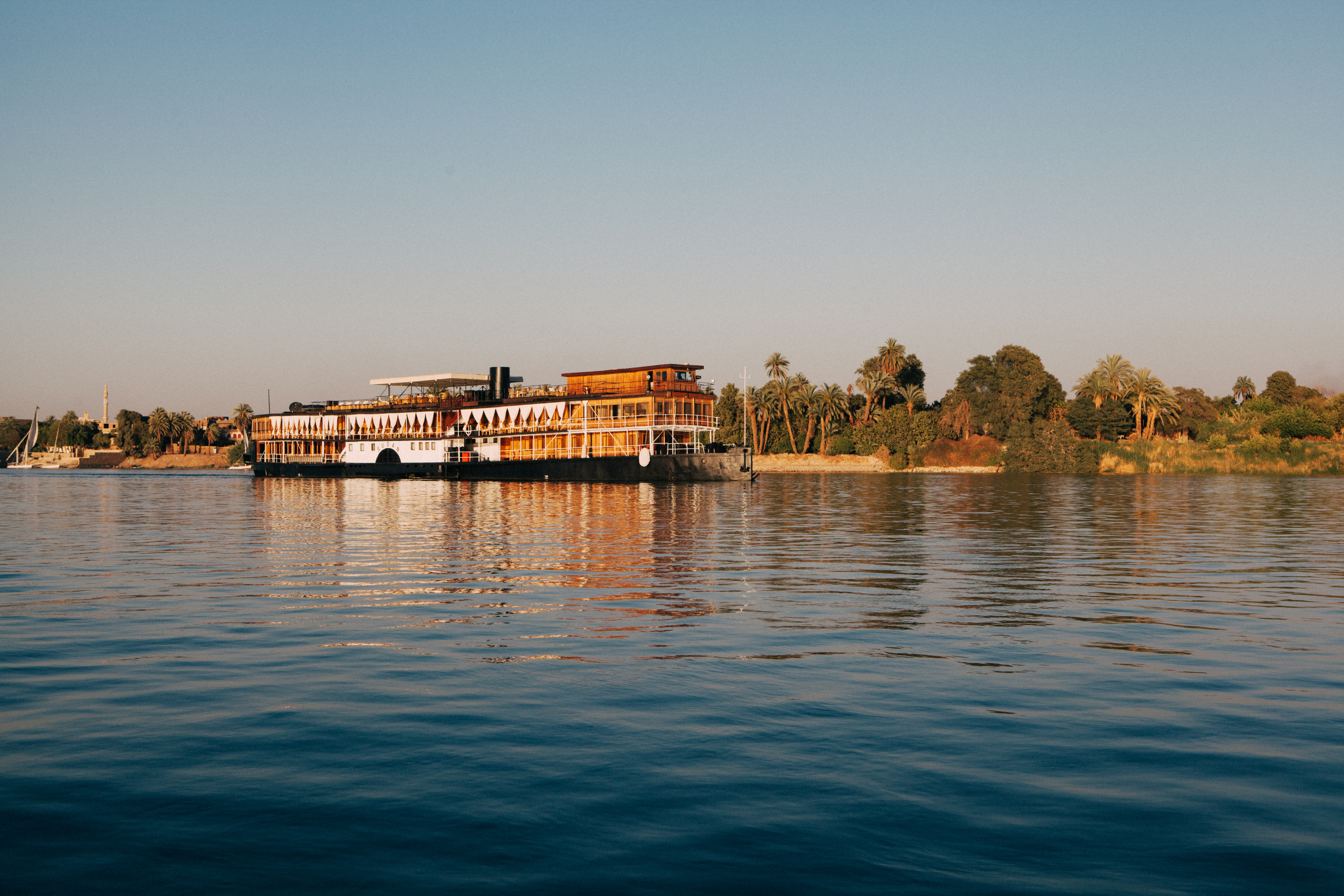Riverfront building and a moored boat sit on calm water, with gentle ripples and shimmering reflections in warm light.