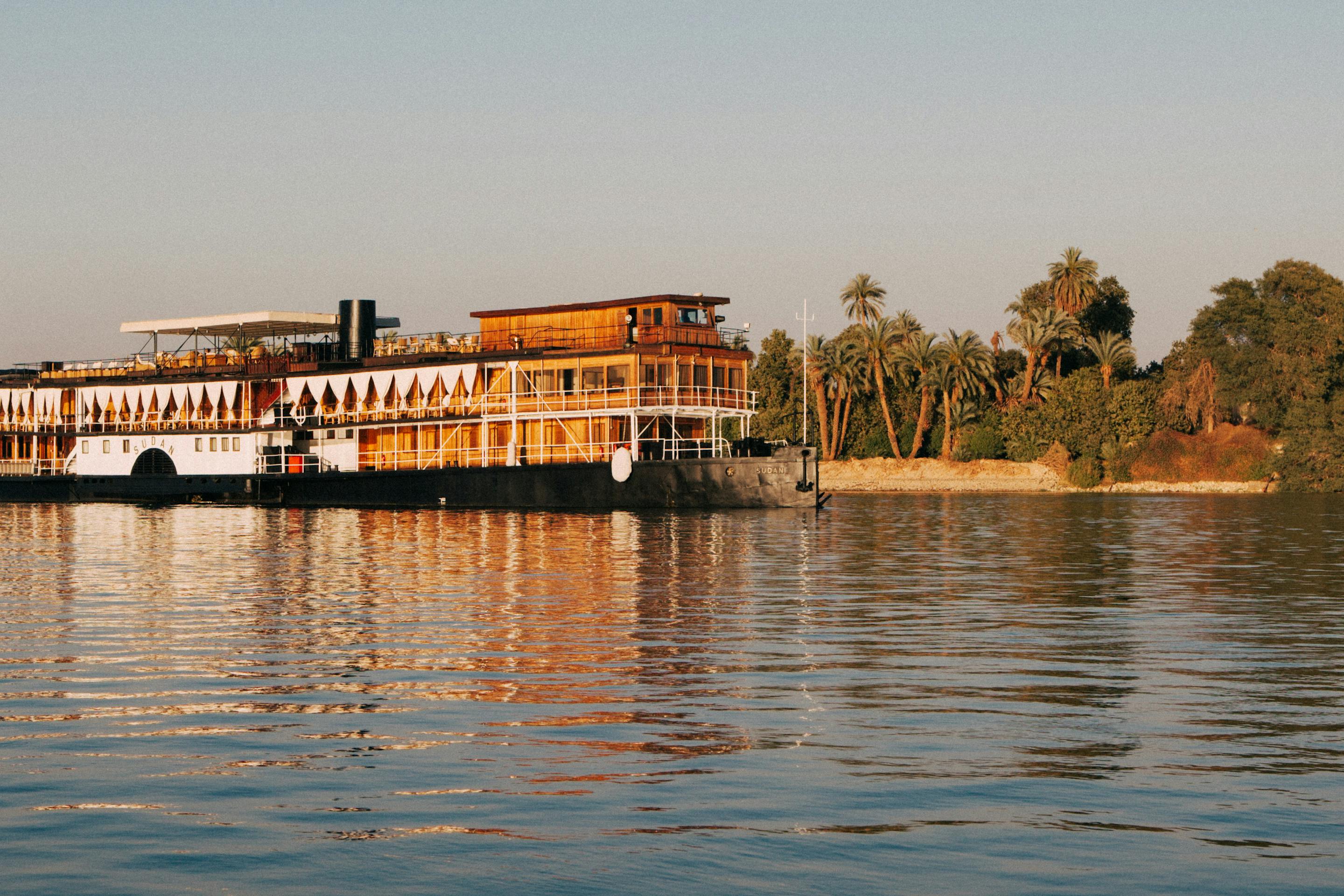Riverfront building and a moored boat sit on calm water, with gentle ripples and shimmering reflections in warm light.