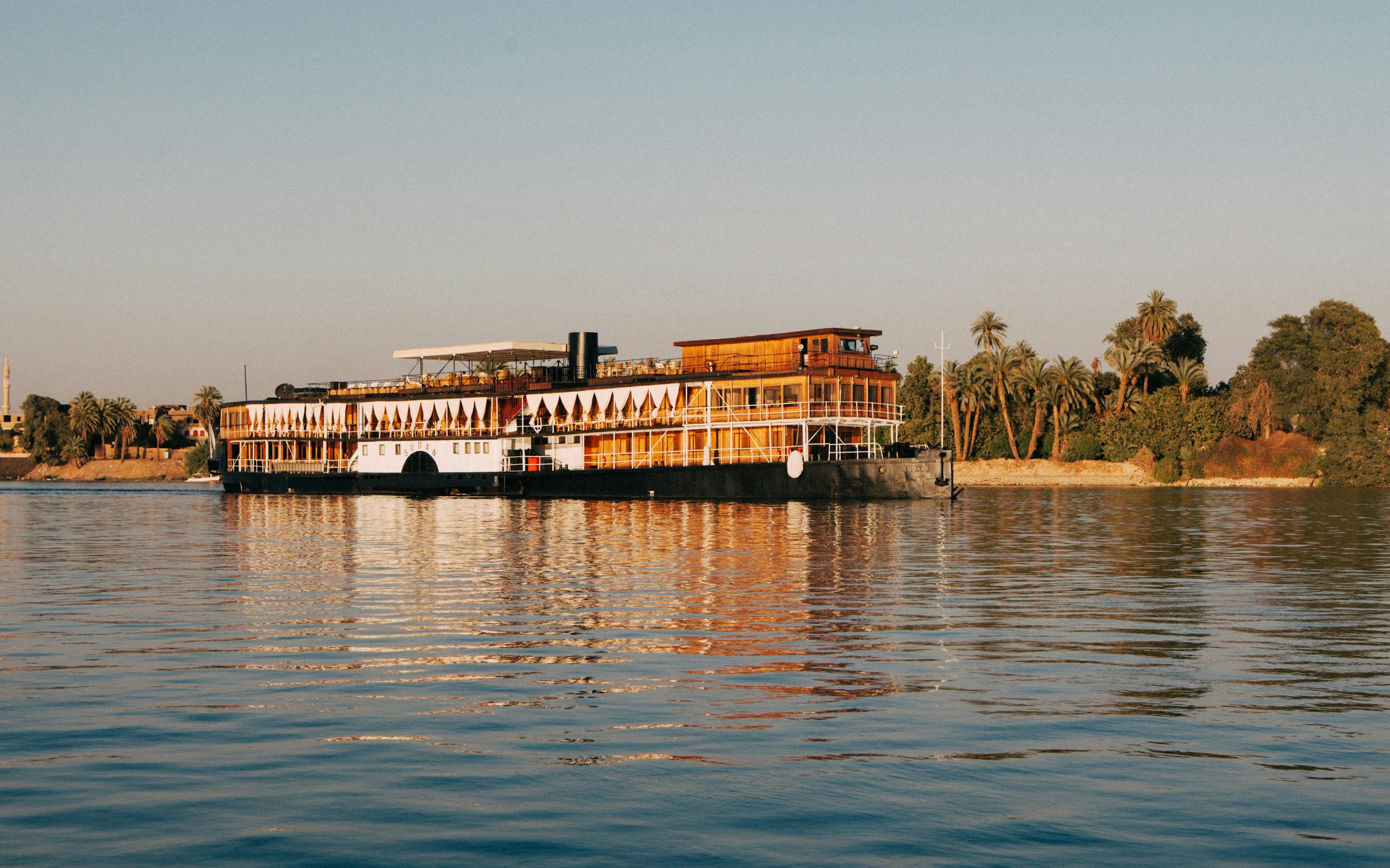Riverfront building and a moored boat sit on calm water, with gentle ripples and shimmering reflections in warm light.