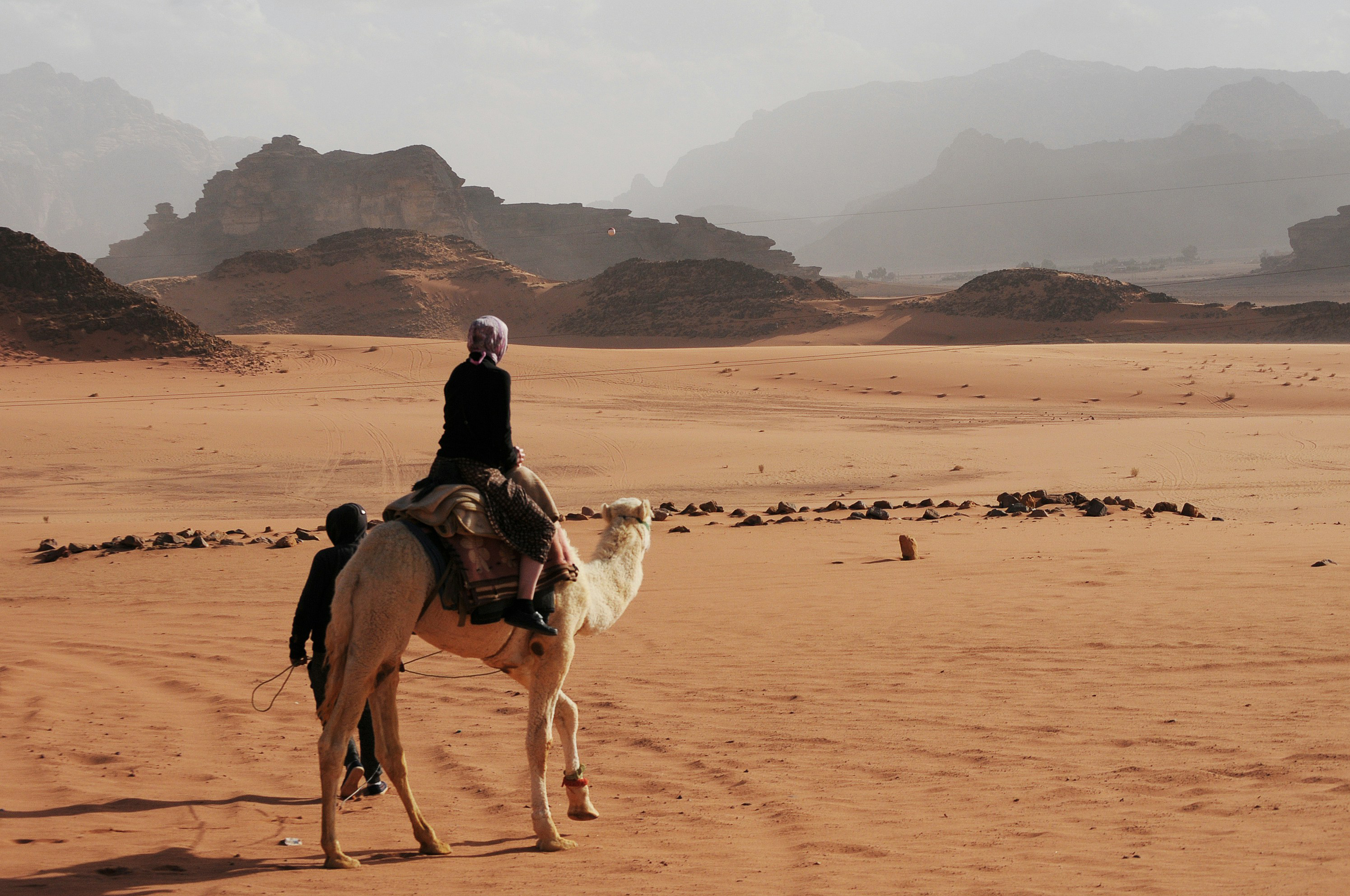 Camel with a rider crosses a sandy desert plain, leaving tracks toward distant rock formations under soft clouds.