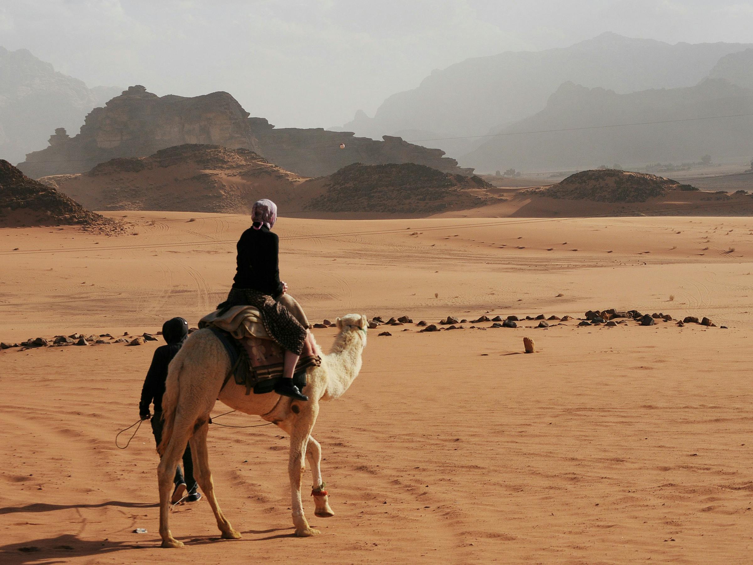 Camel with a rider crosses a sandy desert plain, leaving tracks toward distant rock formations under soft clouds.