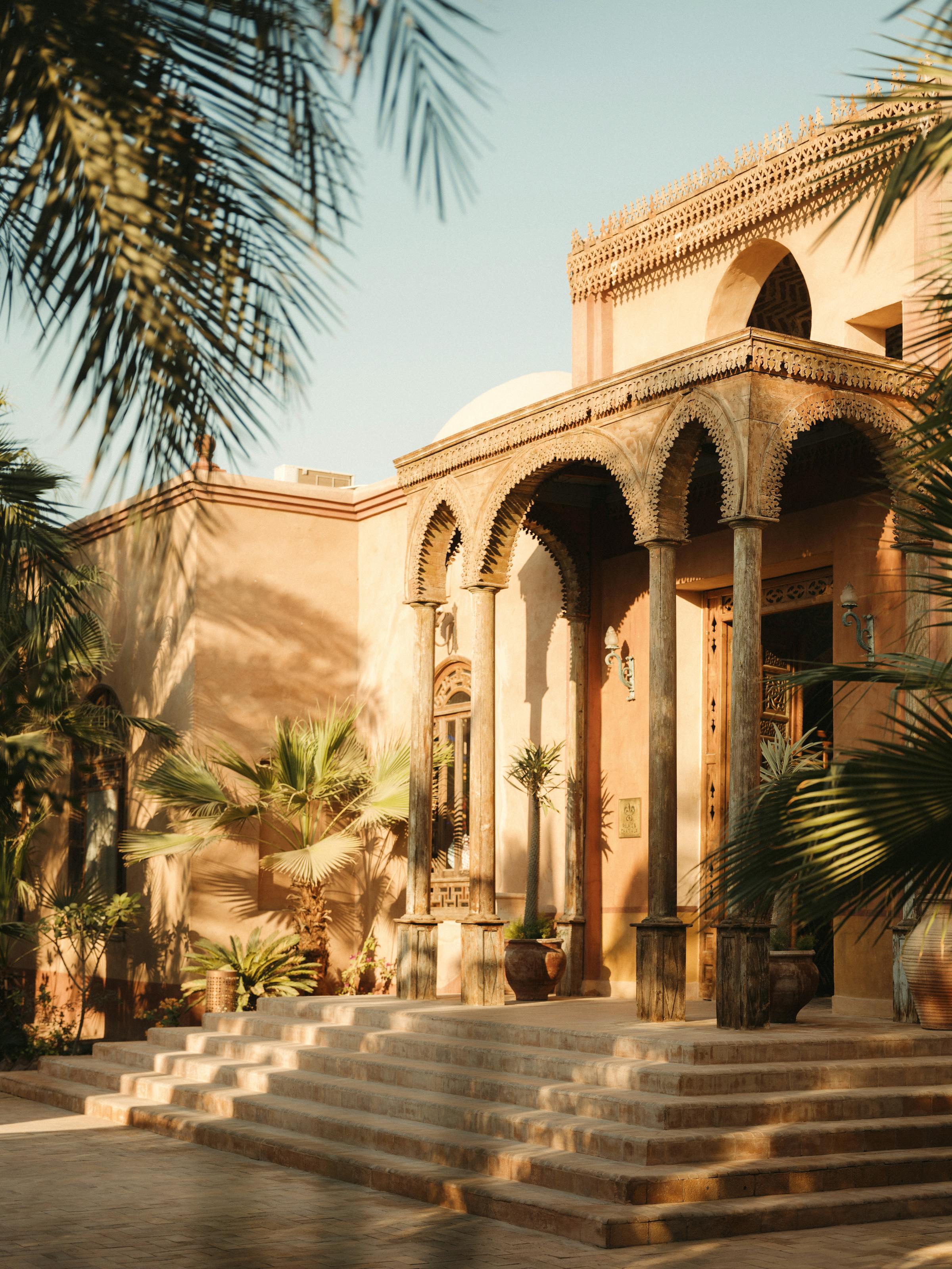 Sunlit hotel entrance with arched columns and steps, framed by palms and long shadows across the walkway.