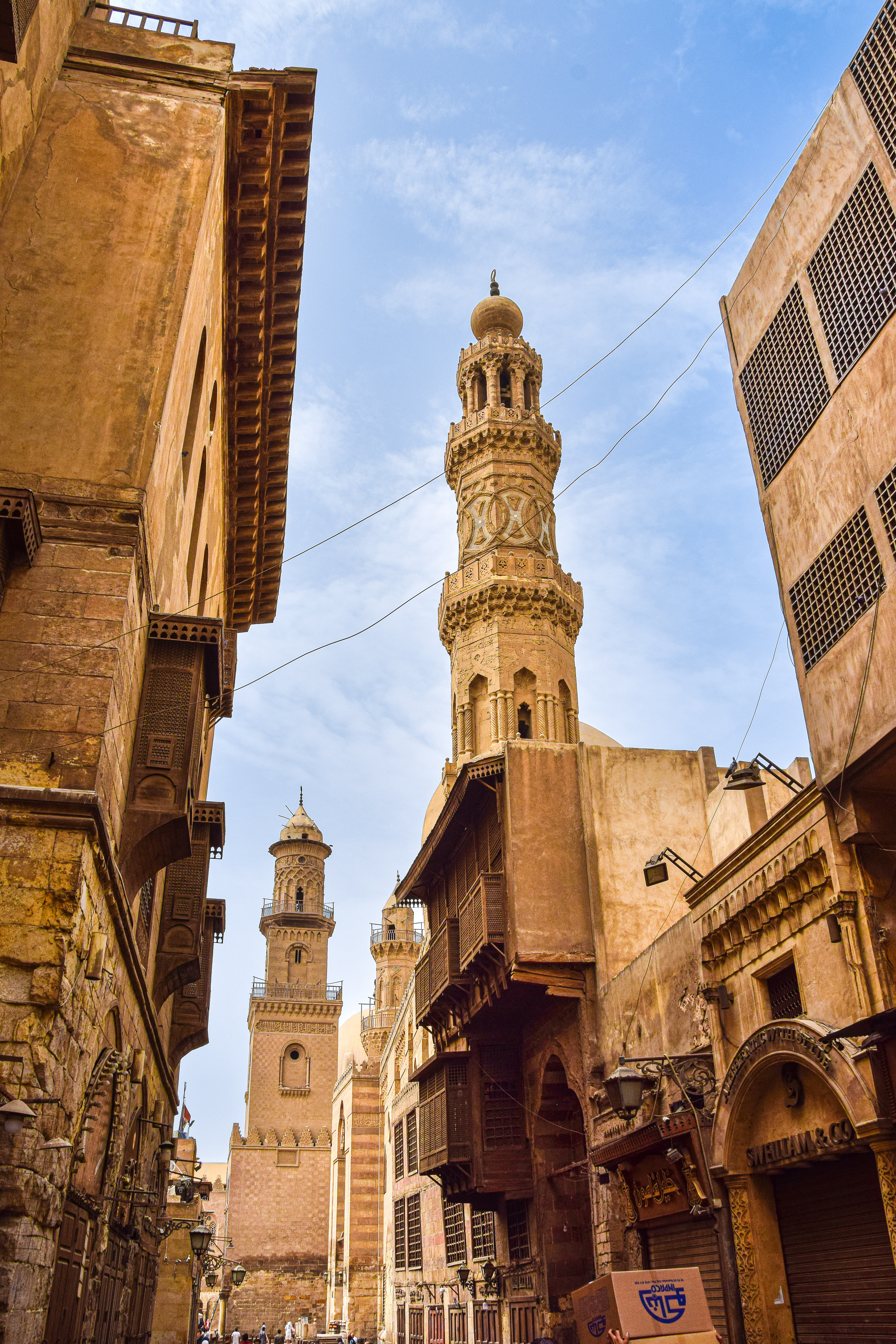 Tall minaret tower rises above a narrow street, with old stone buildings and blue sky between the rooftops.