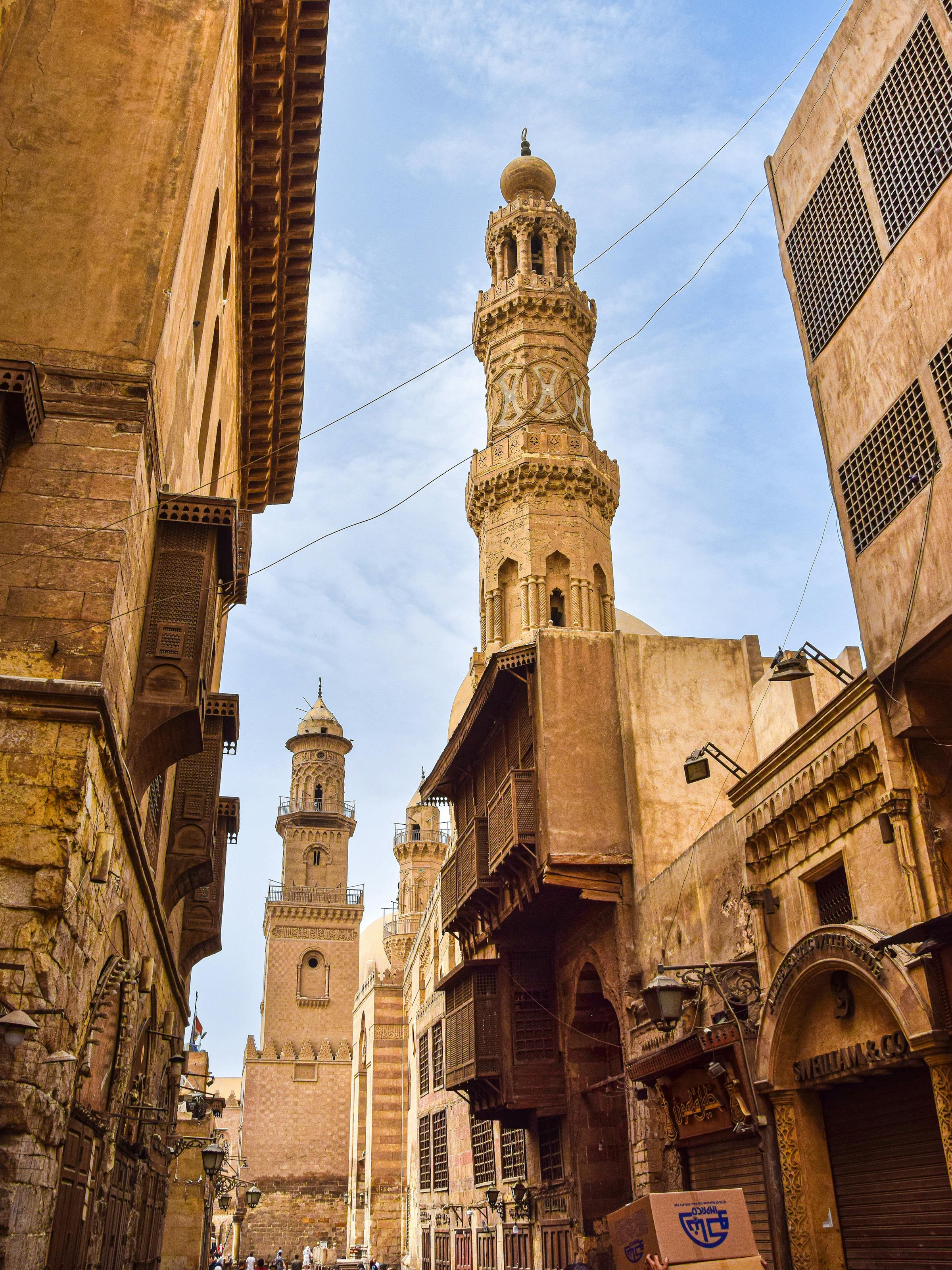Tall minaret tower rises above a narrow street, with old stone buildings and blue sky between the rooftops.